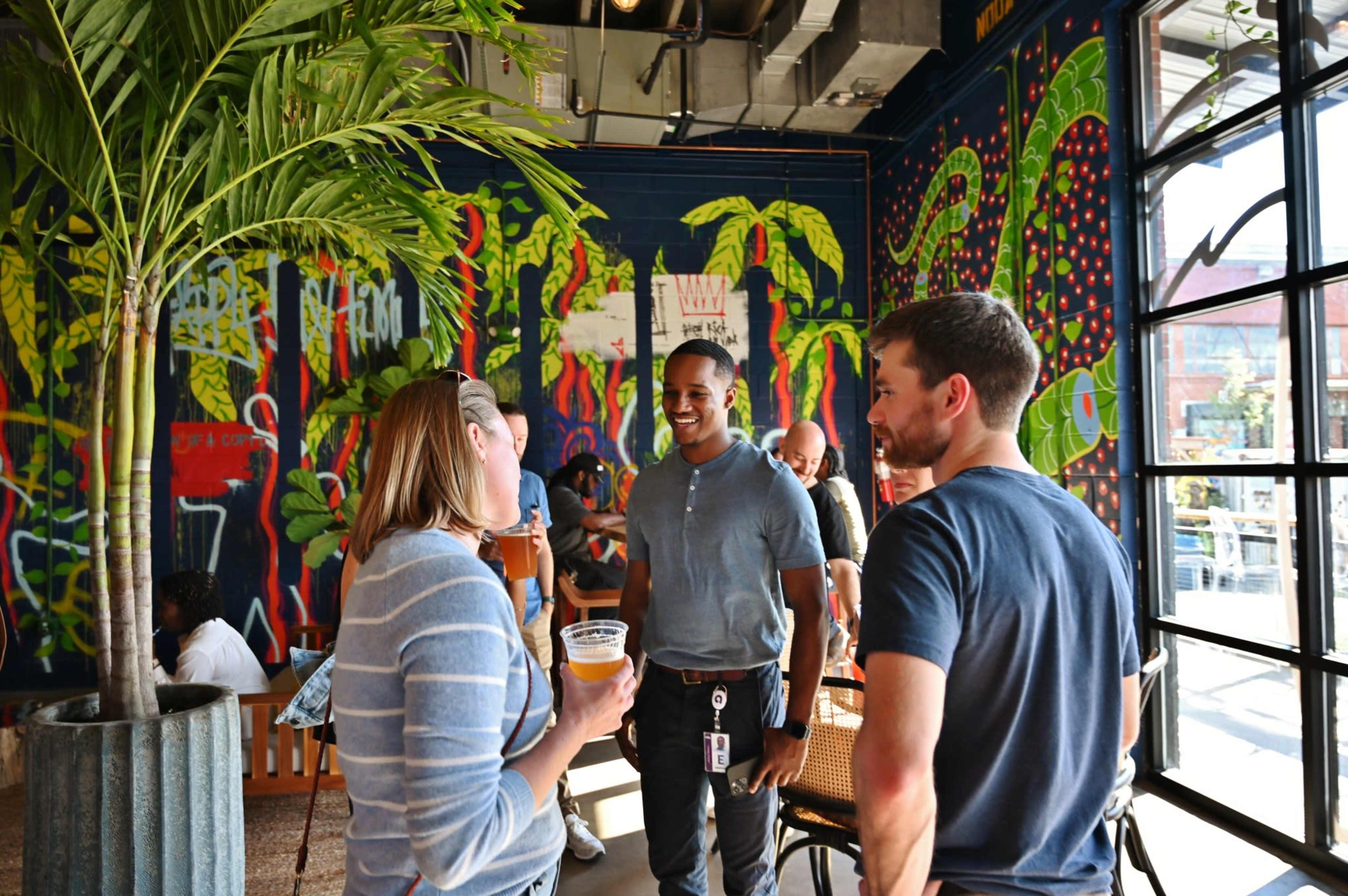 A group of people engage in conversation inside a venue decorated with vibrant plant-themed murals.