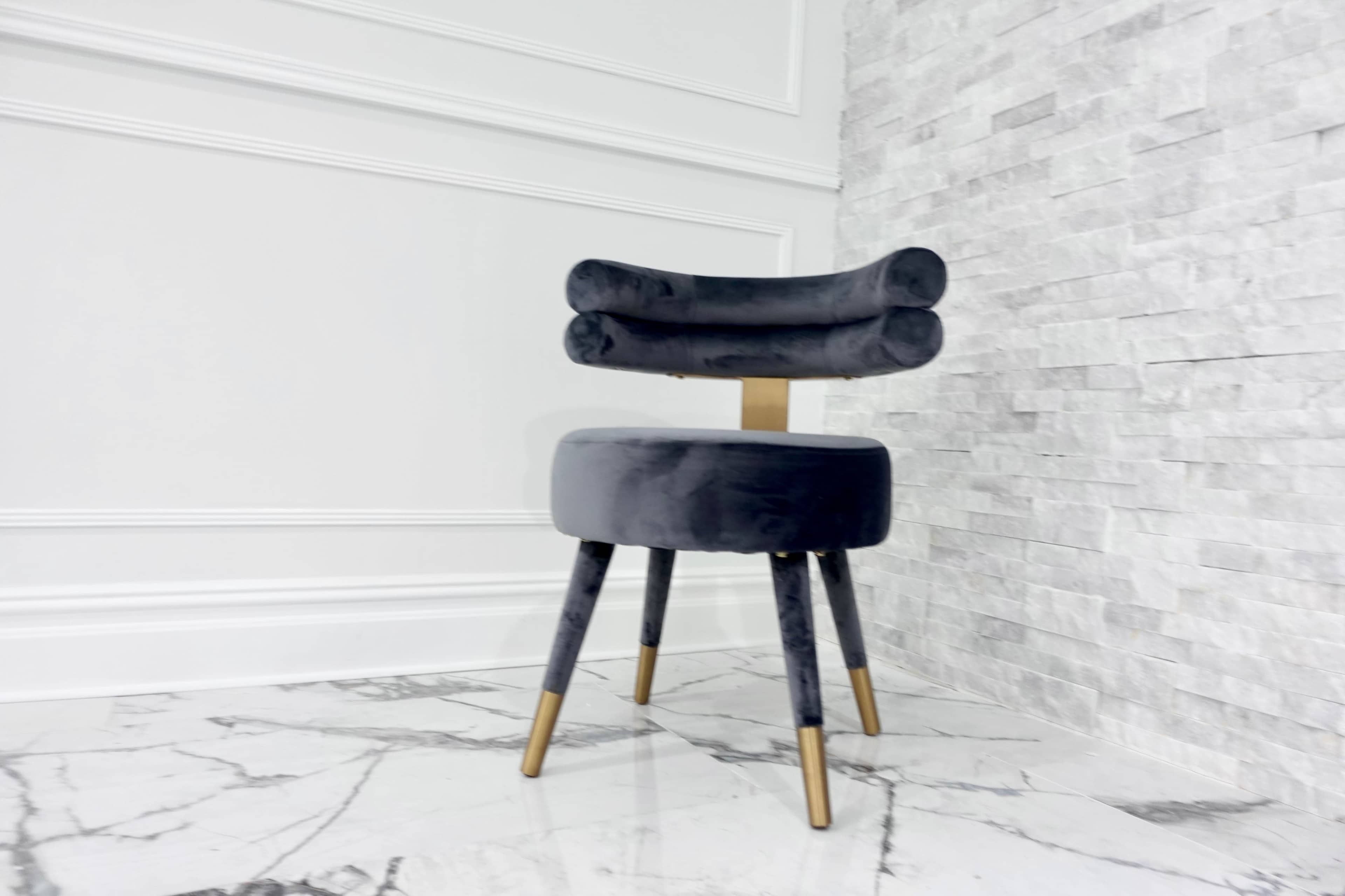 A modern black velvet chair with a unique back design stands against a light stone wall.