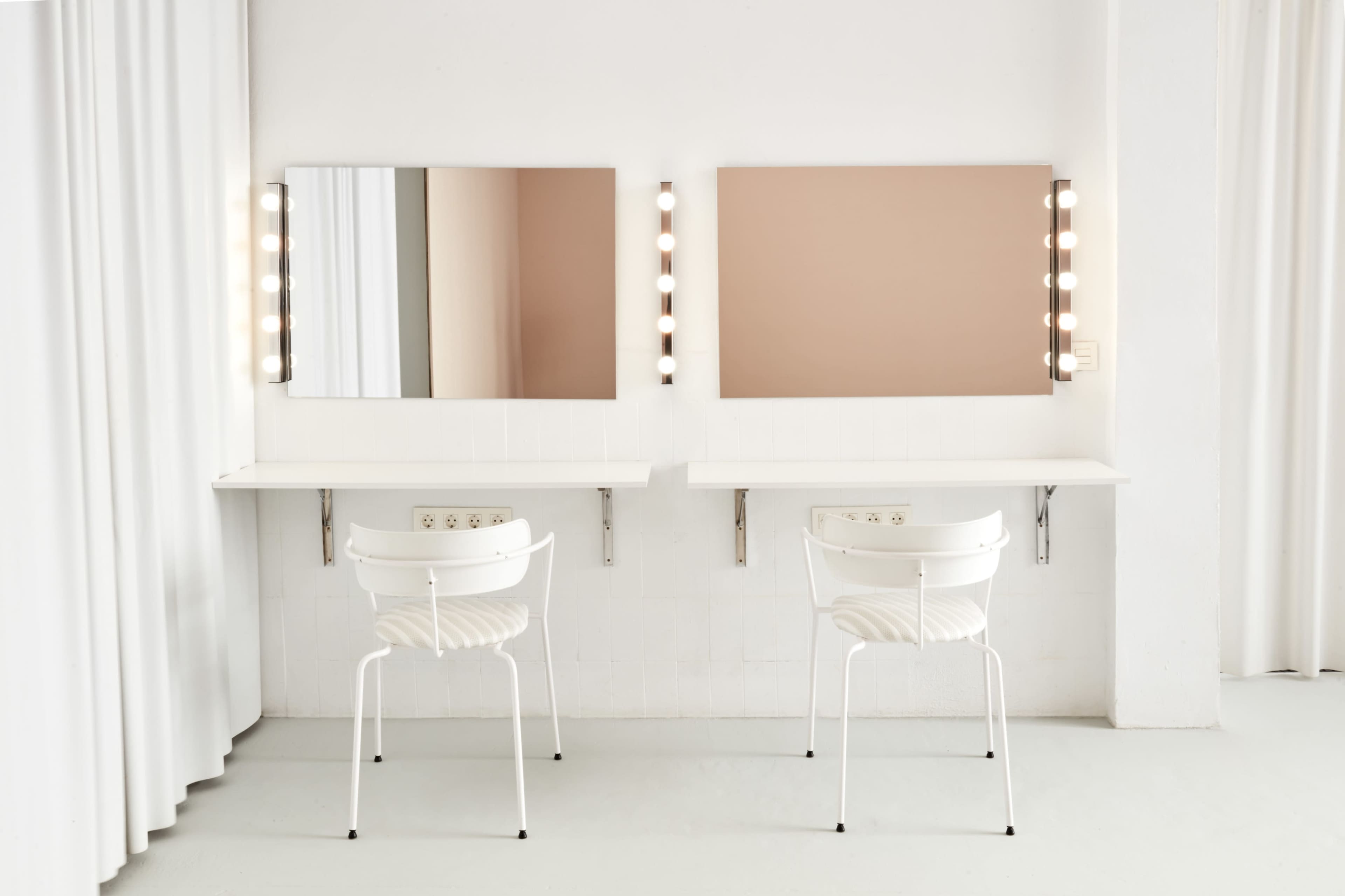 The image shows two white chairs facing two mirrors mounted on a light-colored wall with illuminated borders.