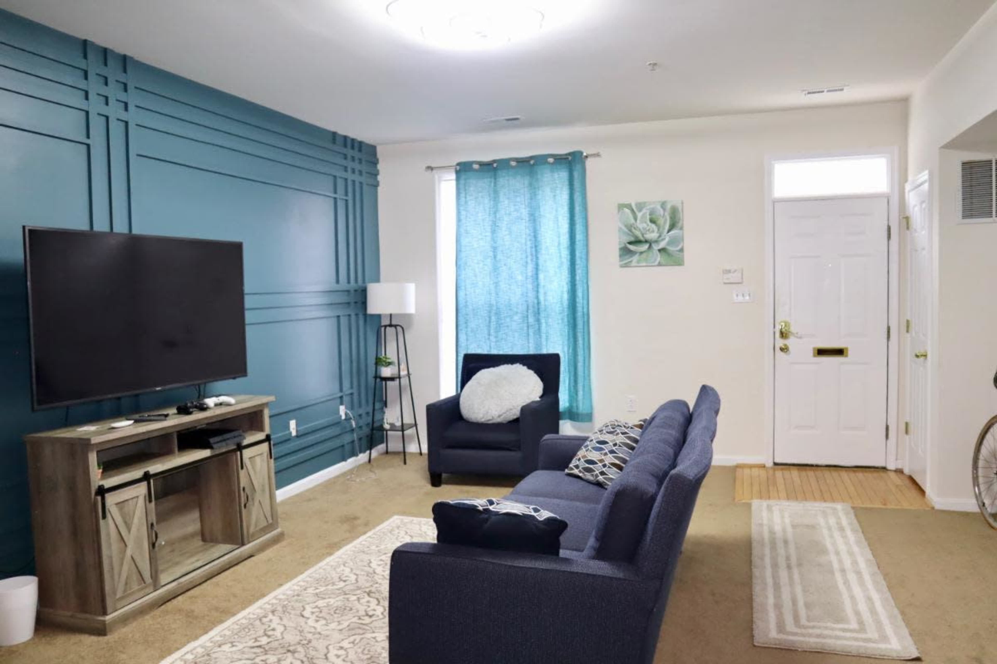 A living room with a blue accent wall, a dark sofa facing a TV, a light-colored area rug, and a window with teal curtains.
