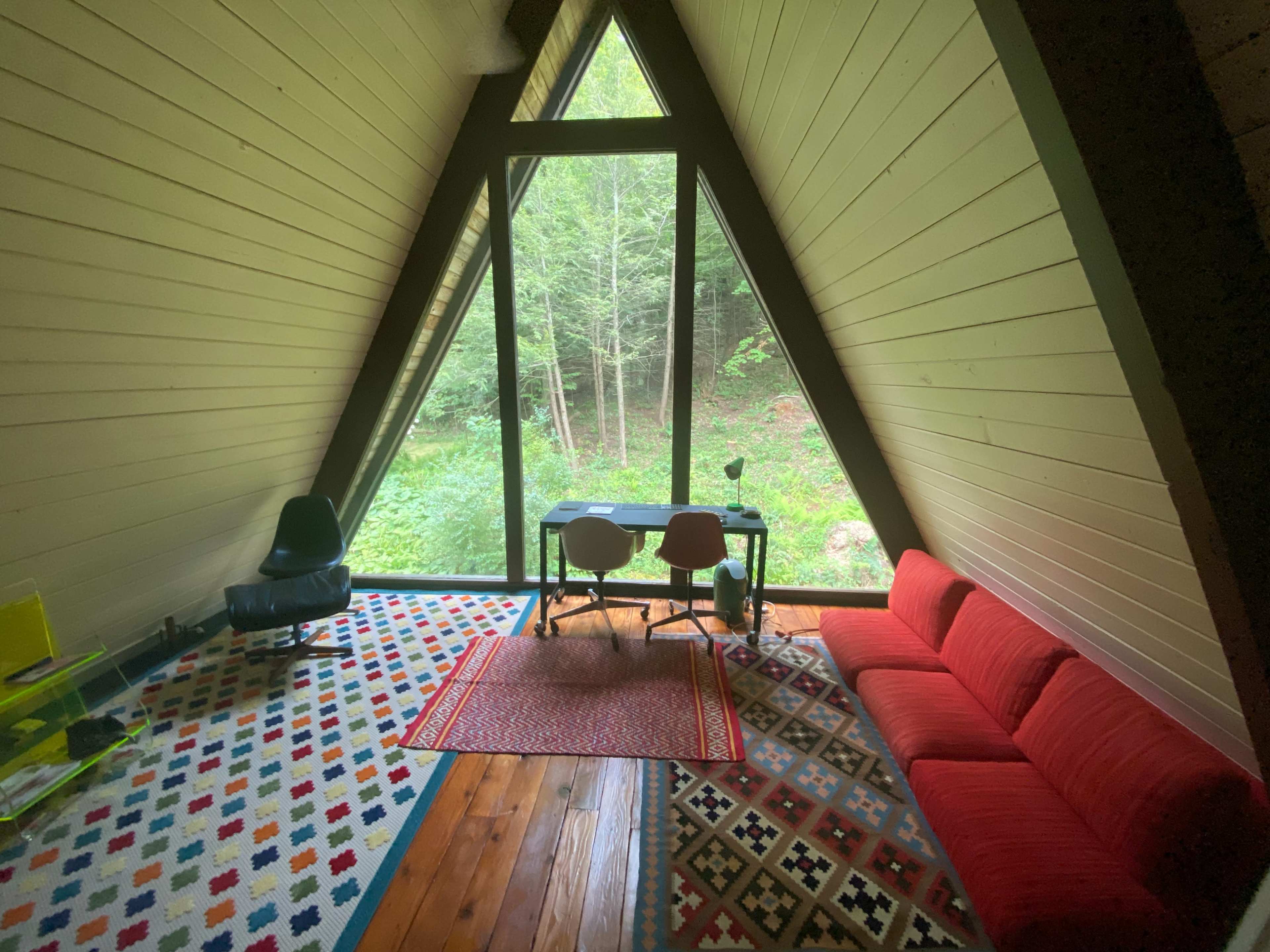 An A-frame interior with a red couch, a black chair, and a desk positioned before large windows facing a green forest.