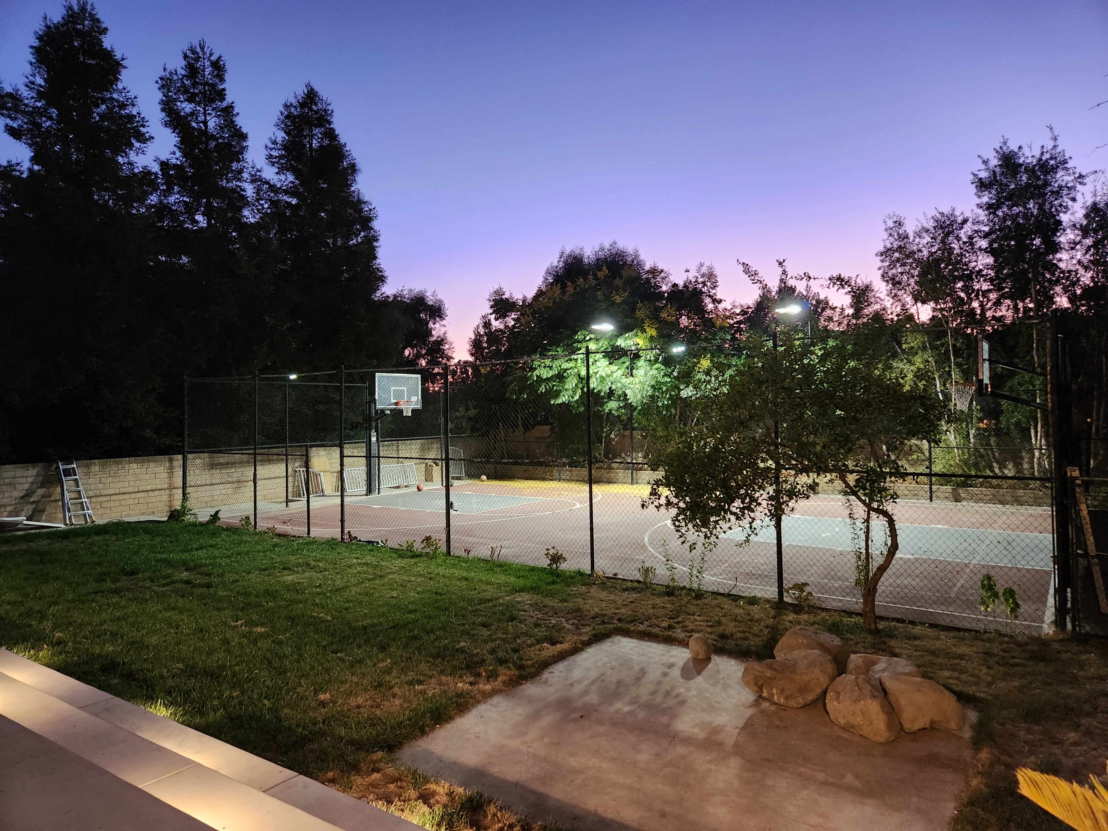 A lit outdoor basketball court is visible at dusk, surrounded by trees and greenery.