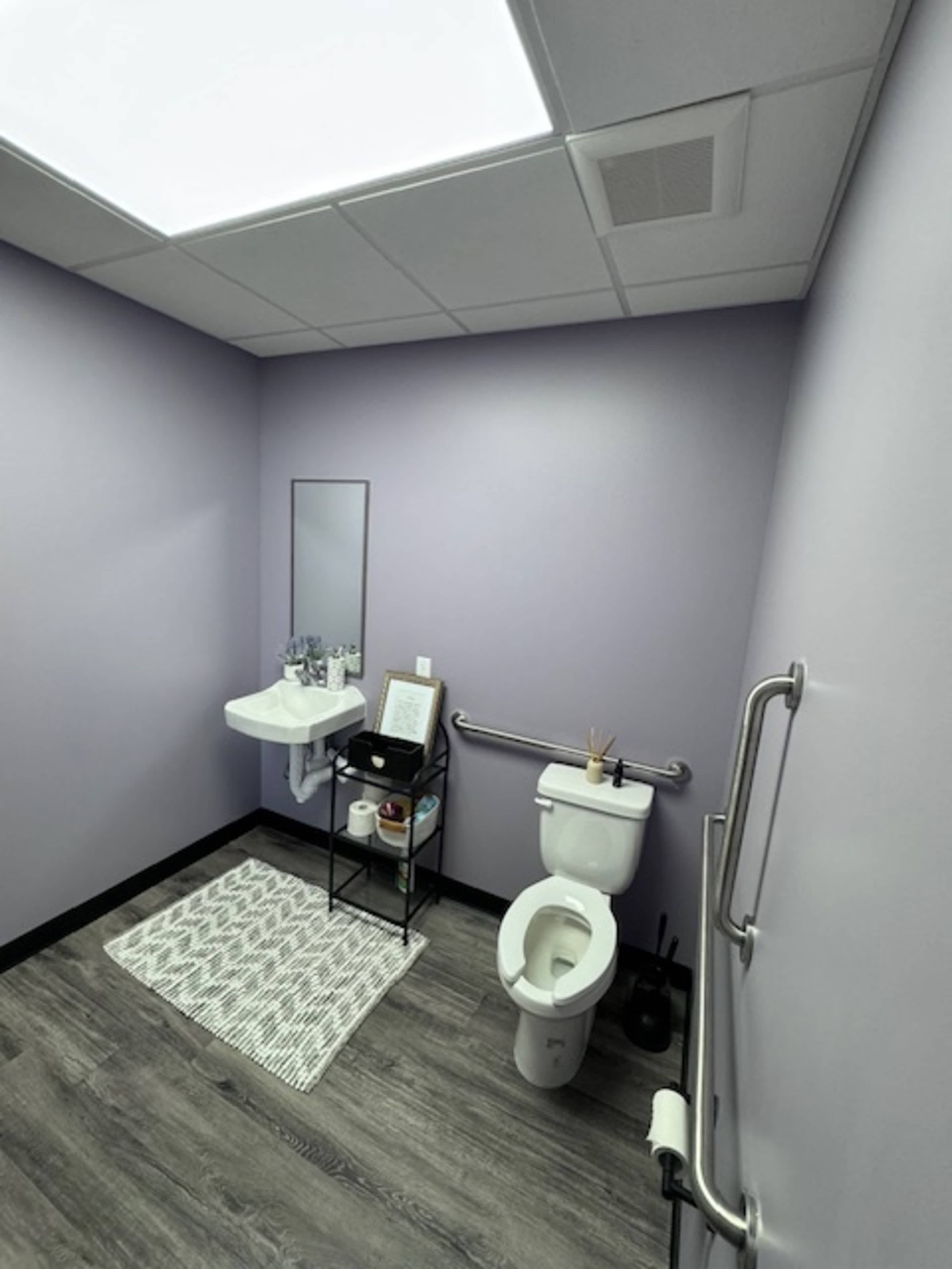 A small, clean restroom featuring a toilet, a sink with a mirror, and a black cart with toiletries against a light purple wall.