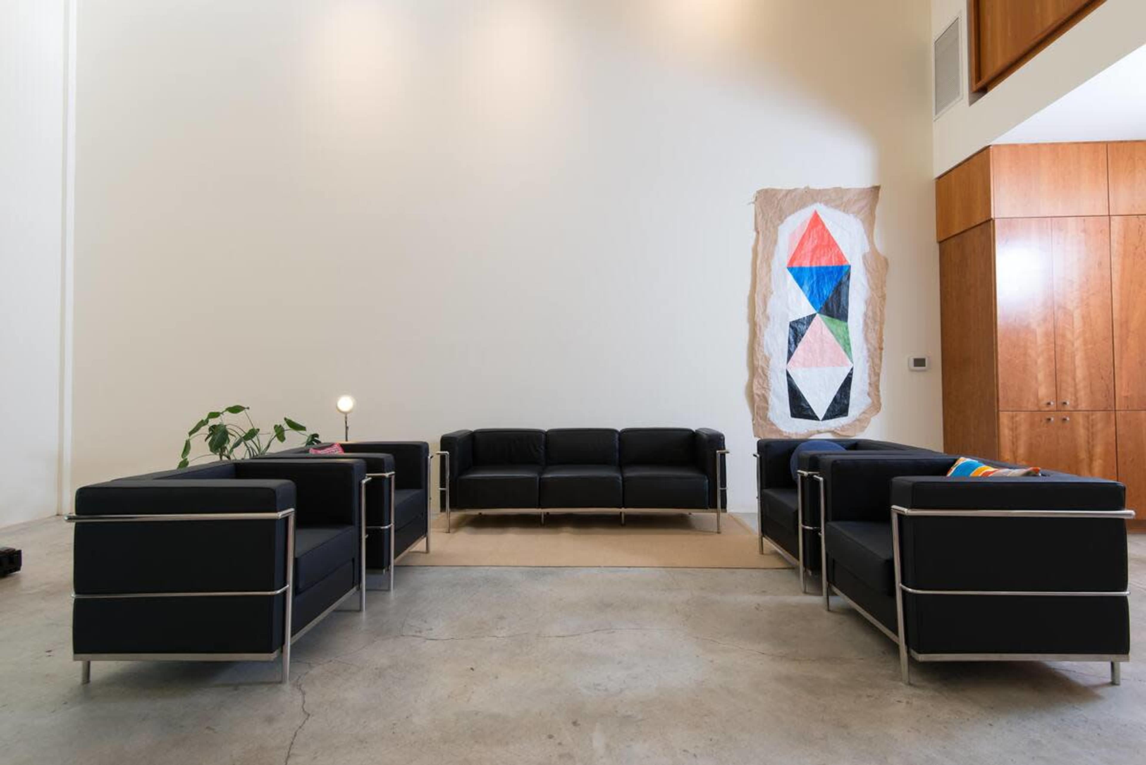 A modern living area with four black sofas arranged around a central space, a geometric wall hanging, and wood cabinetry in the background.