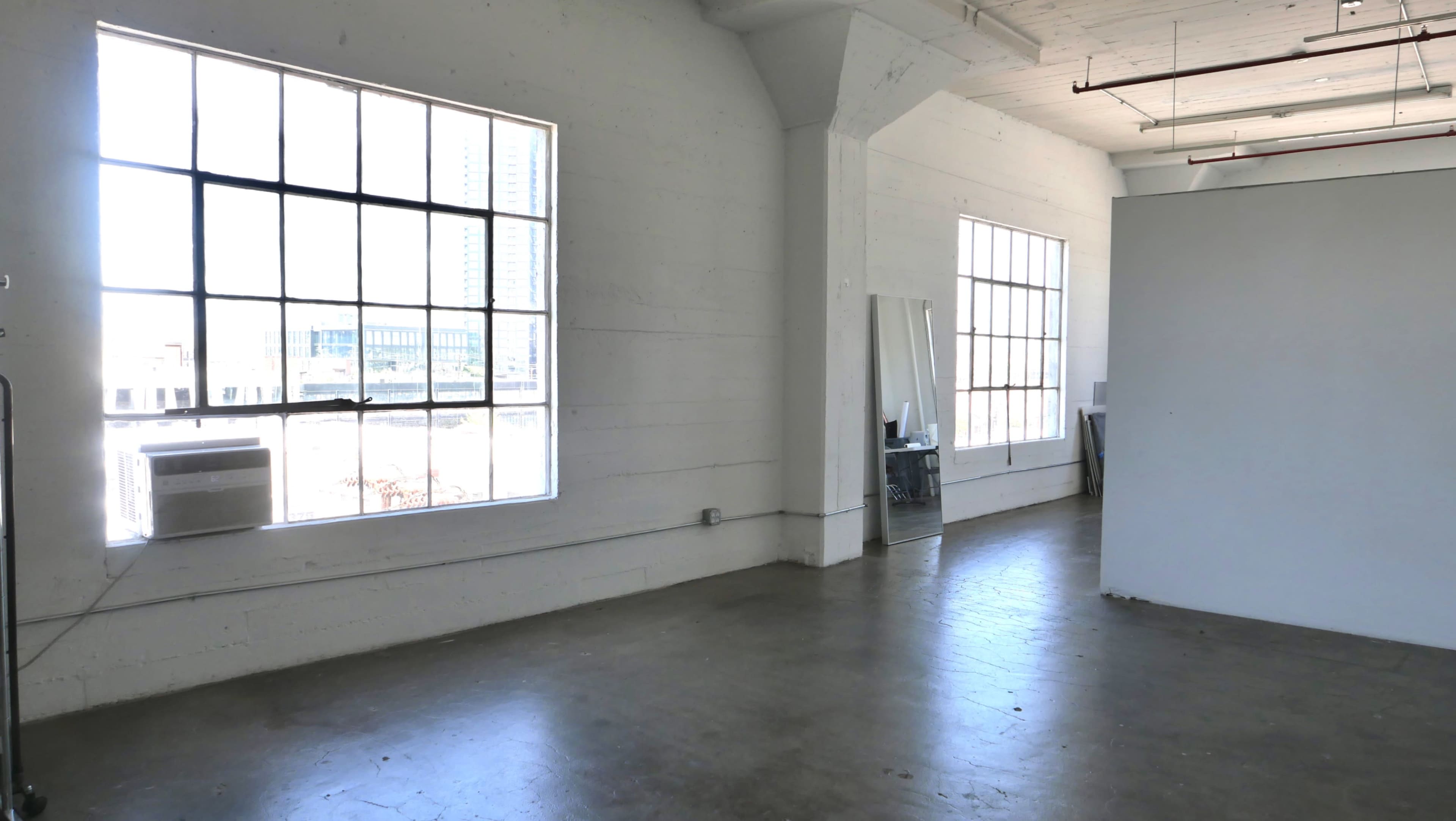 The image shows a spacious, brightly lit room with large windows and a concrete floor, featuring a white partition and a mirror on the wall.