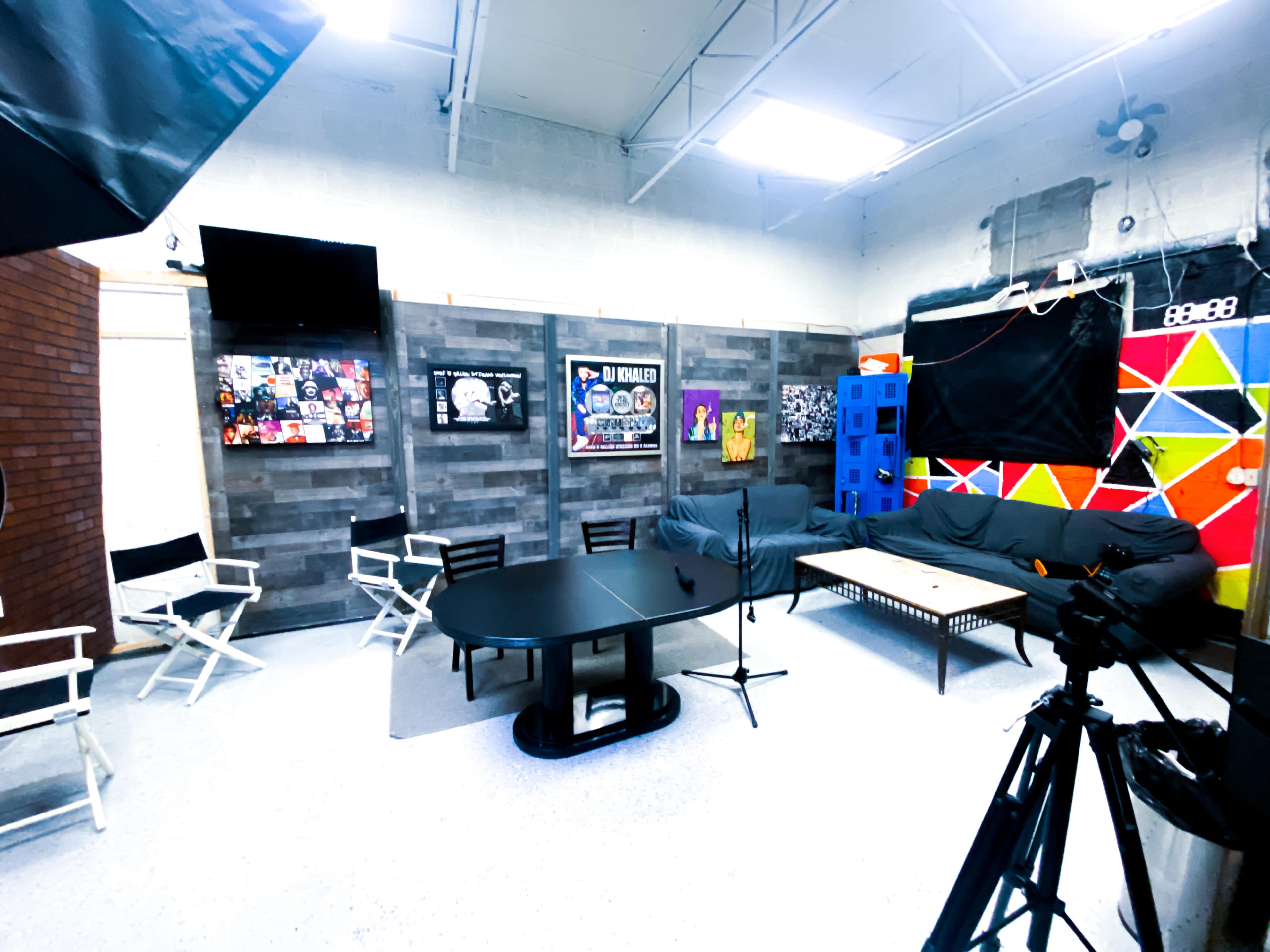 The image shows a studio space with a black table surrounded by chairs, colorful wall decorations, and camera equipment set up for recording.