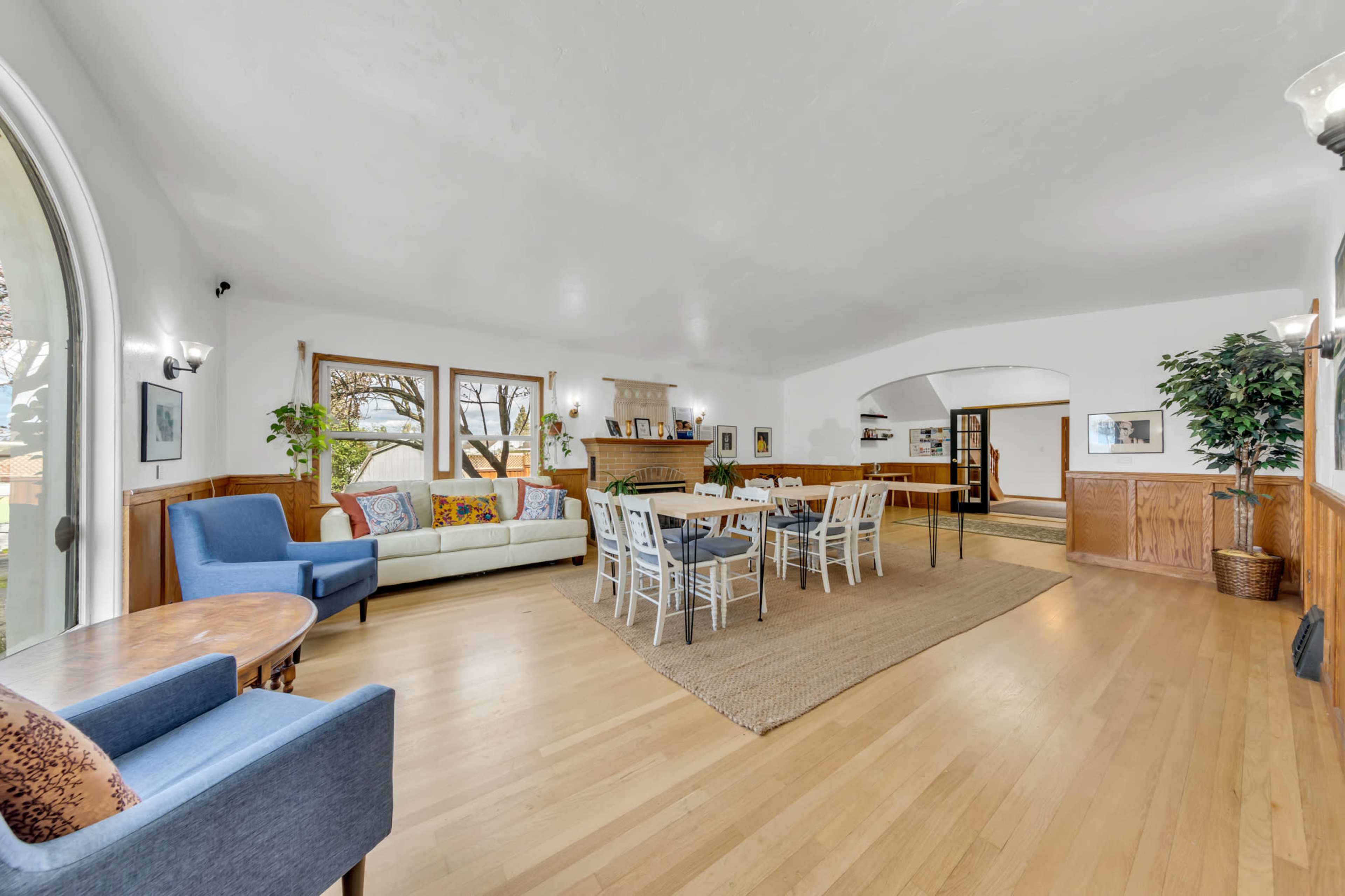 The spacious living area features a light-colored wooden floor, large windows, and a mix of seating arrangements, including a sofa and dining tables.