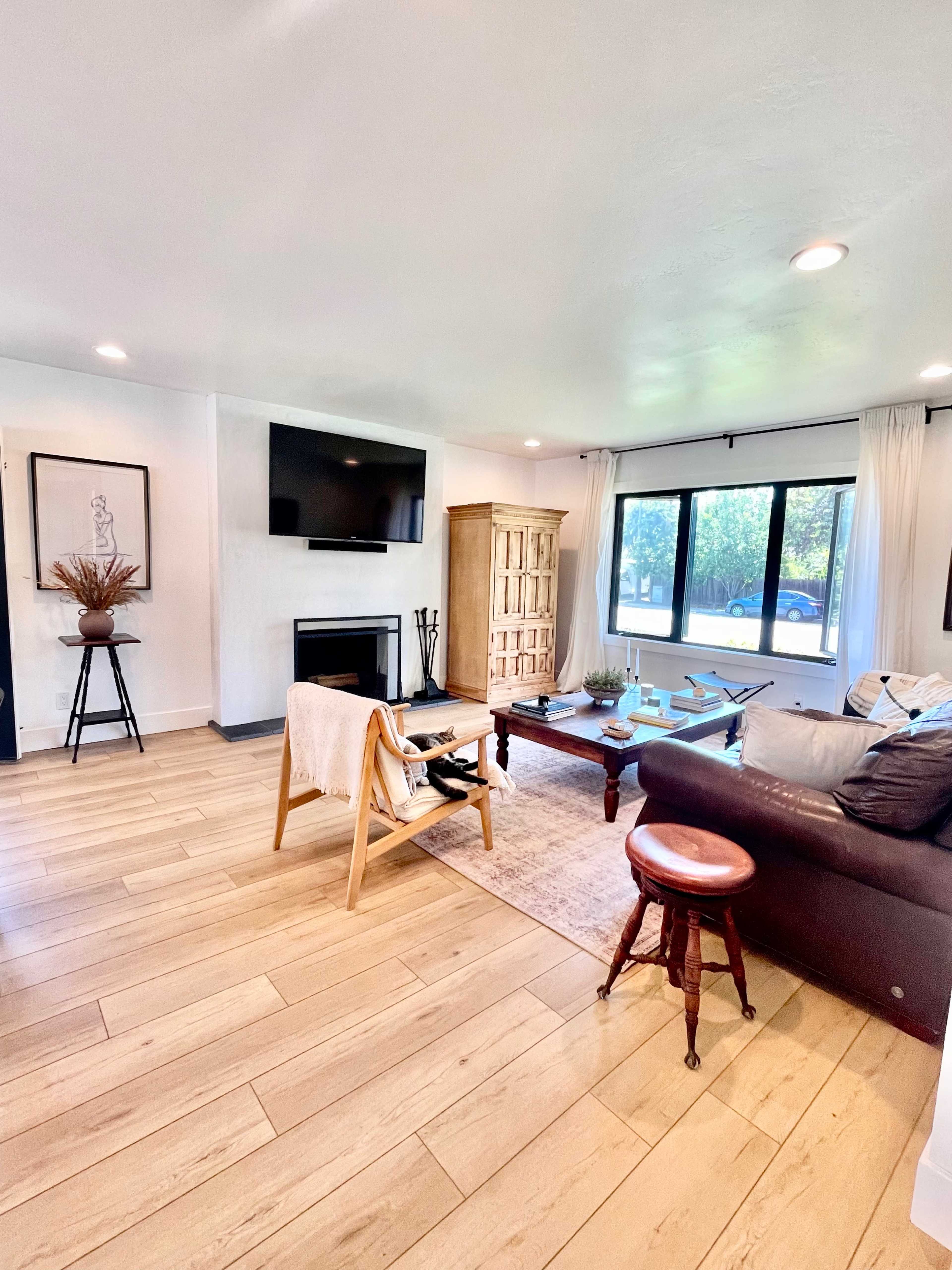 A bright living room with a flat-screen TV mounted on the wall, a wooden cabinet, and a seating area featuring a leather sofa and a wooden table.