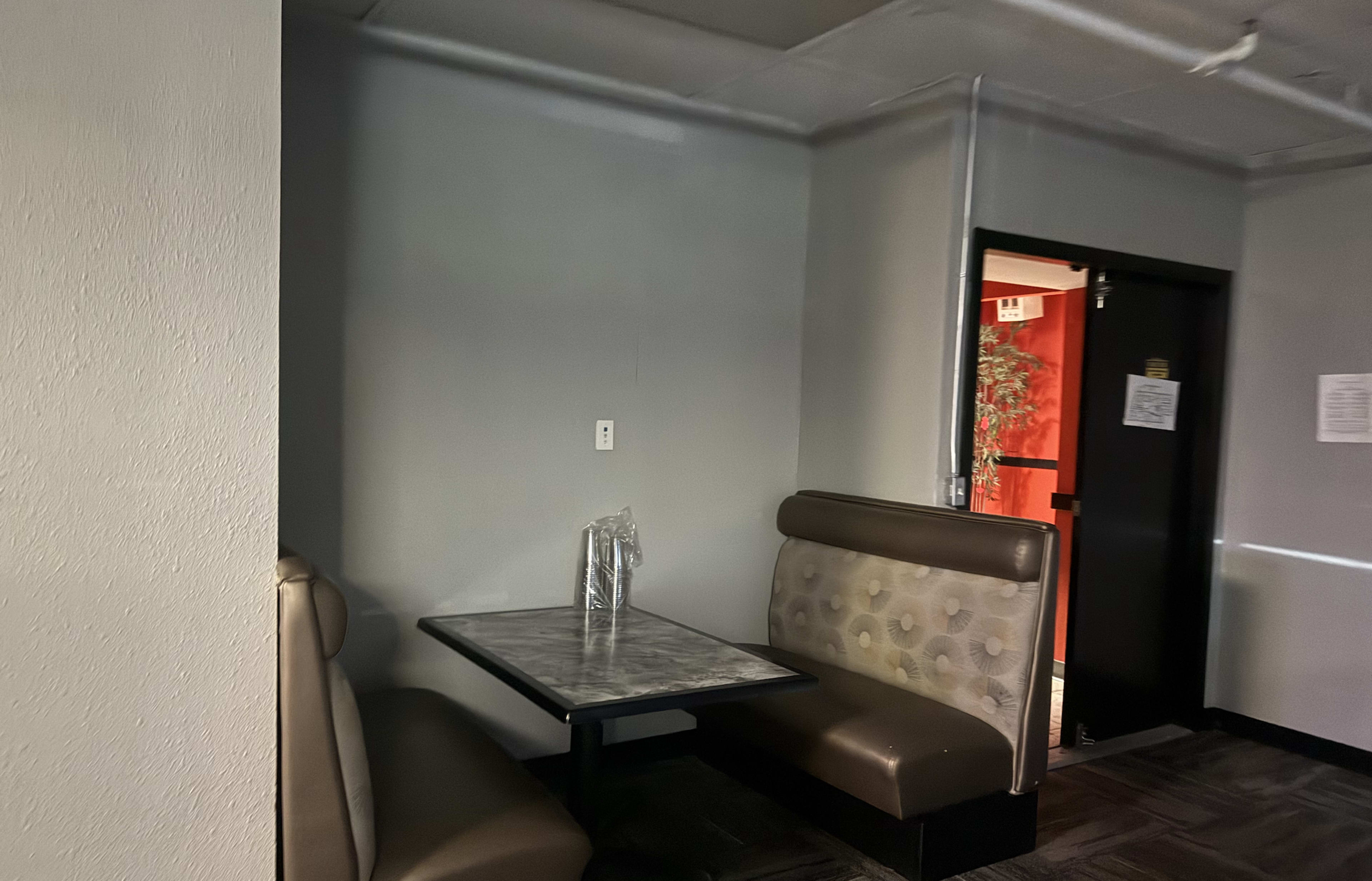 The image shows a small dining area with a booth and a table against a gray wall, near a doorway.