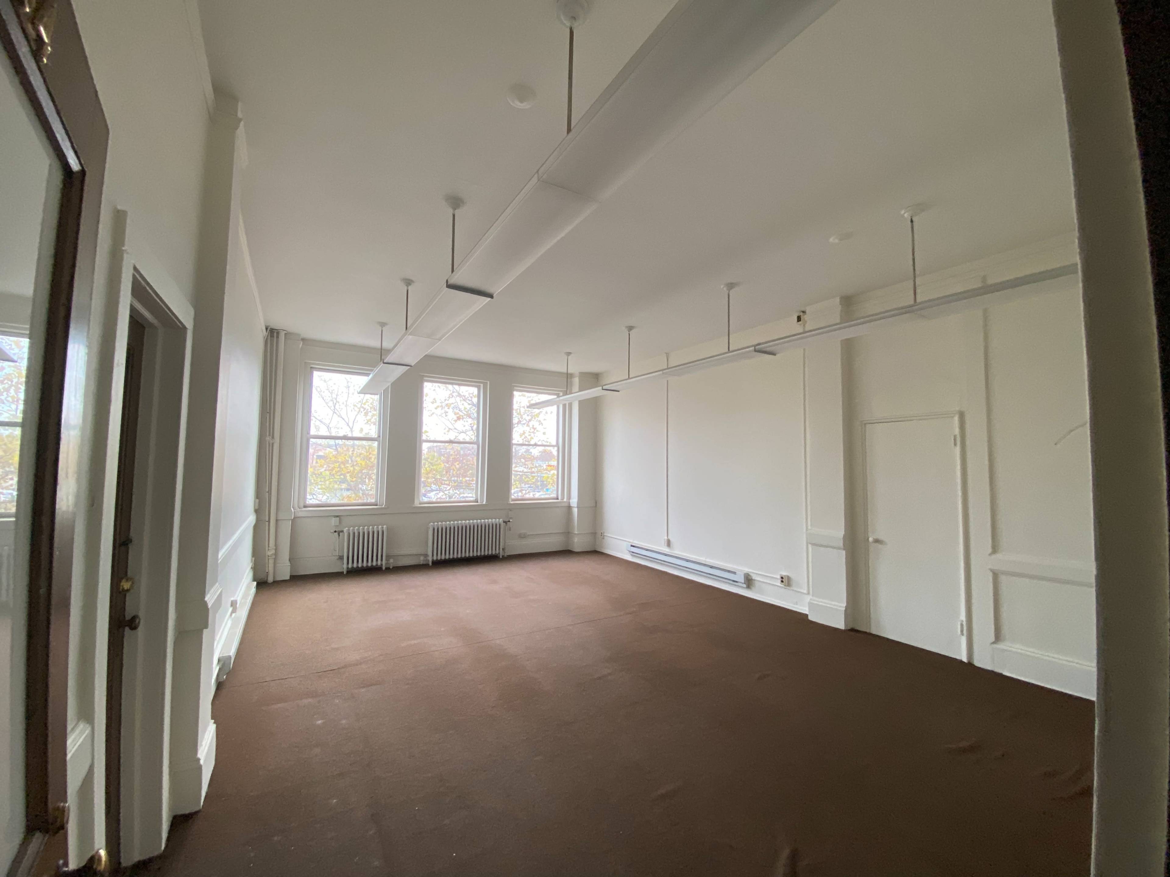 The image shows a vacant, sparsely furnished room with large windows, white walls, and brown carpet.