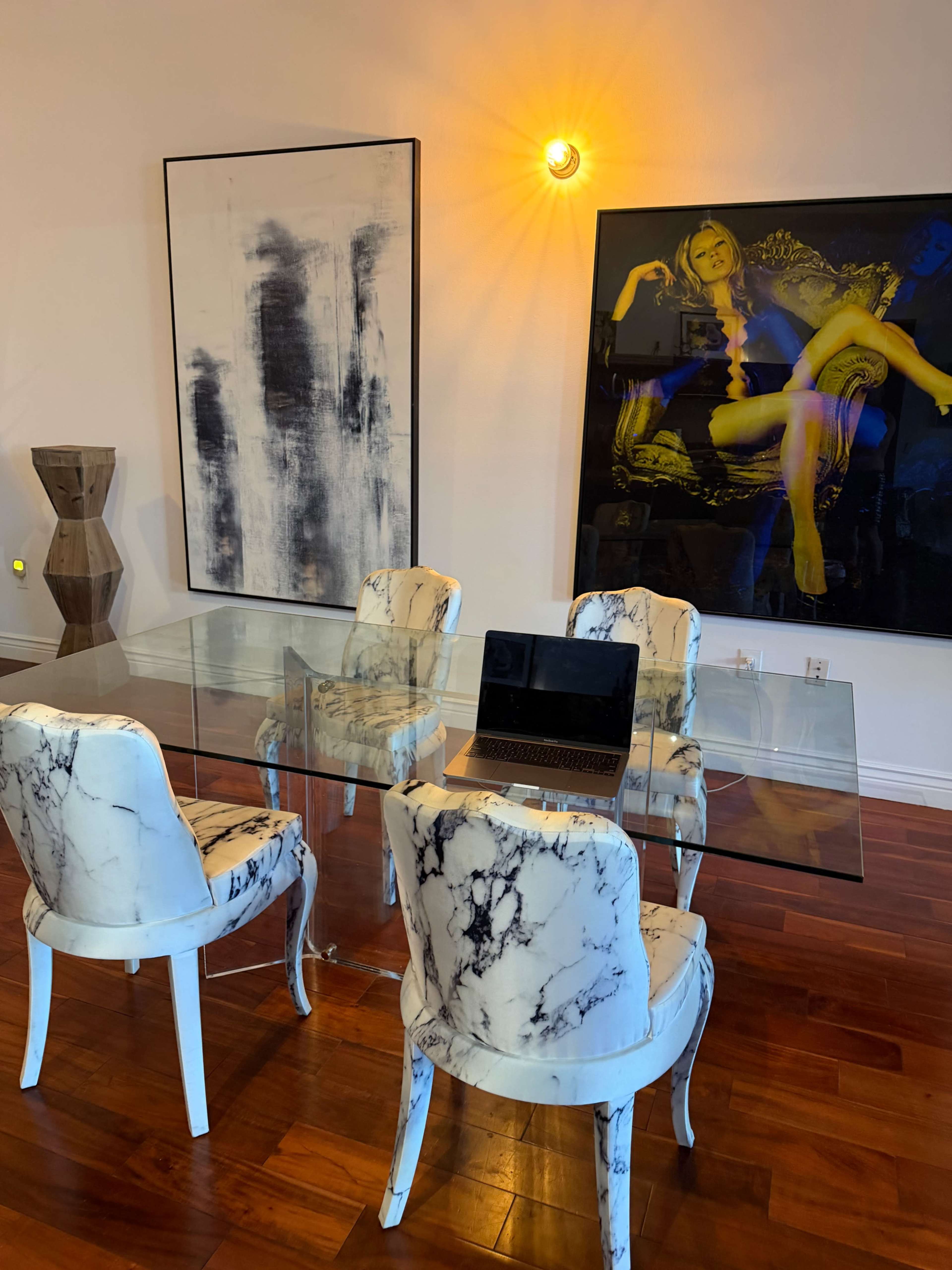 A glass dining table surrounded by four marble-patterned chairs is set against a backdrop of two large artworks on the walls.