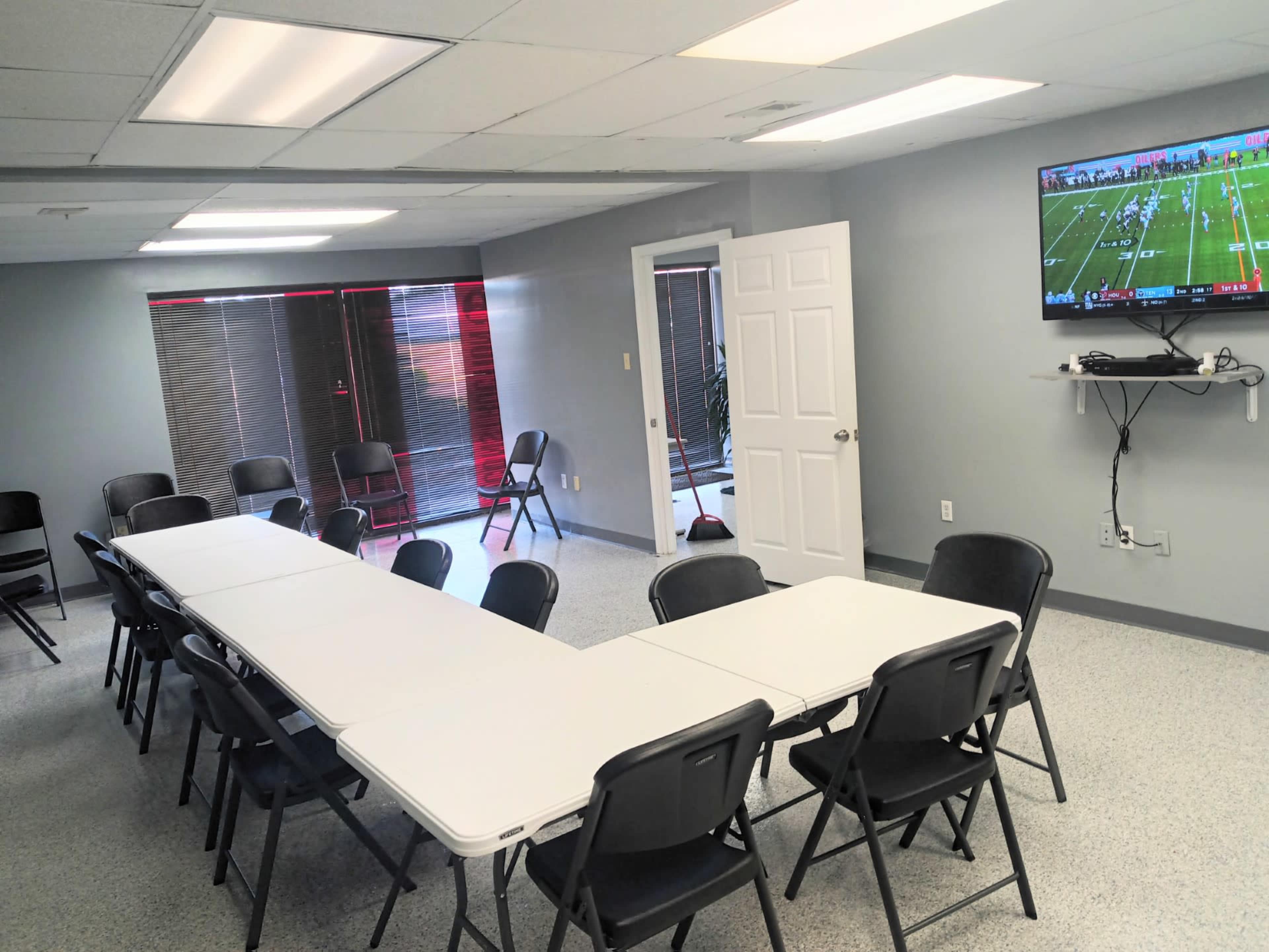 A meeting room with a U-shaped arrangement of white tables surrounded by black chairs and a television mounted on the wall displaying a sports event.