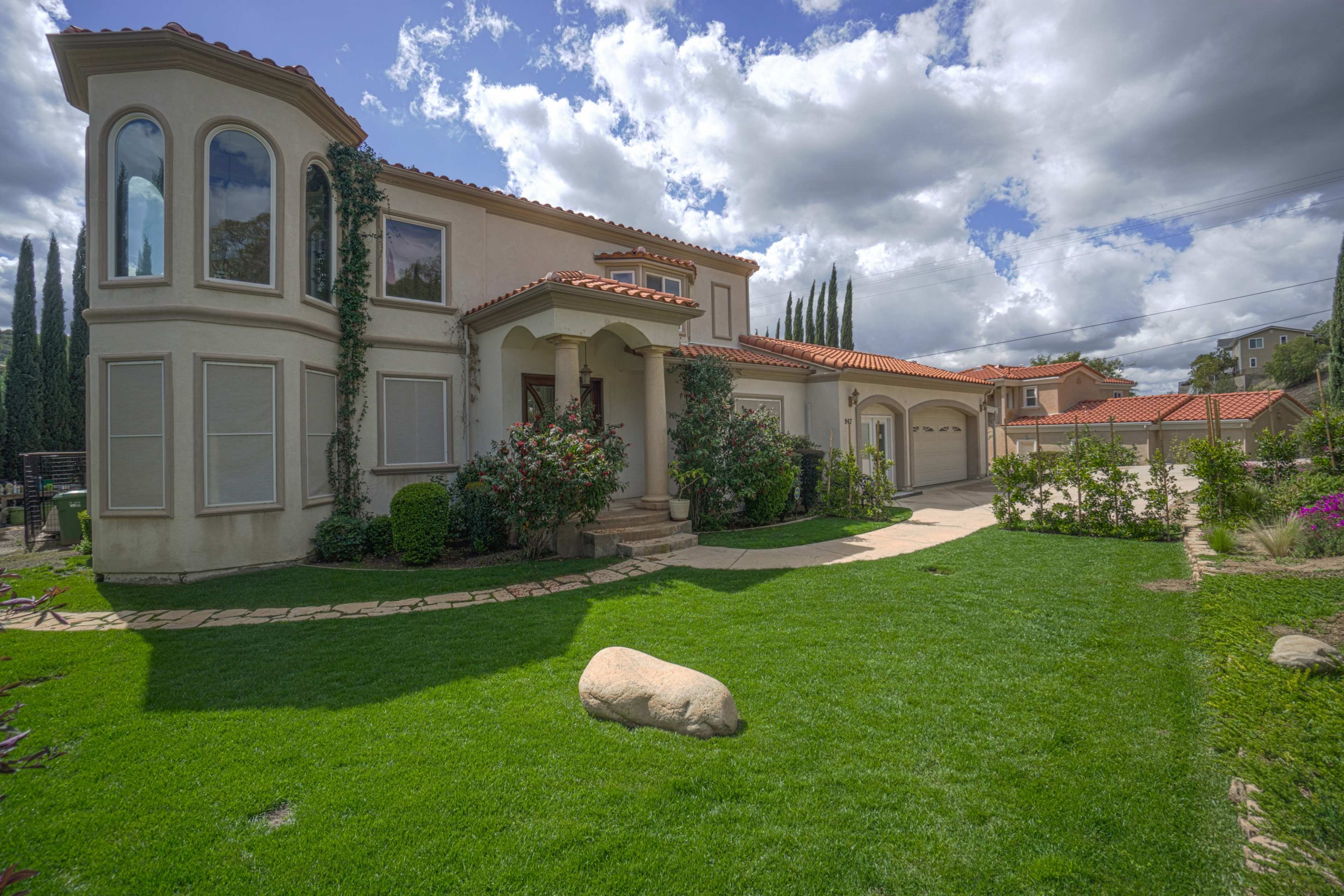 A two-story house with a curved window and a stone pathway is surrounded by green grass and flowering shrubs under a cloudy sky.