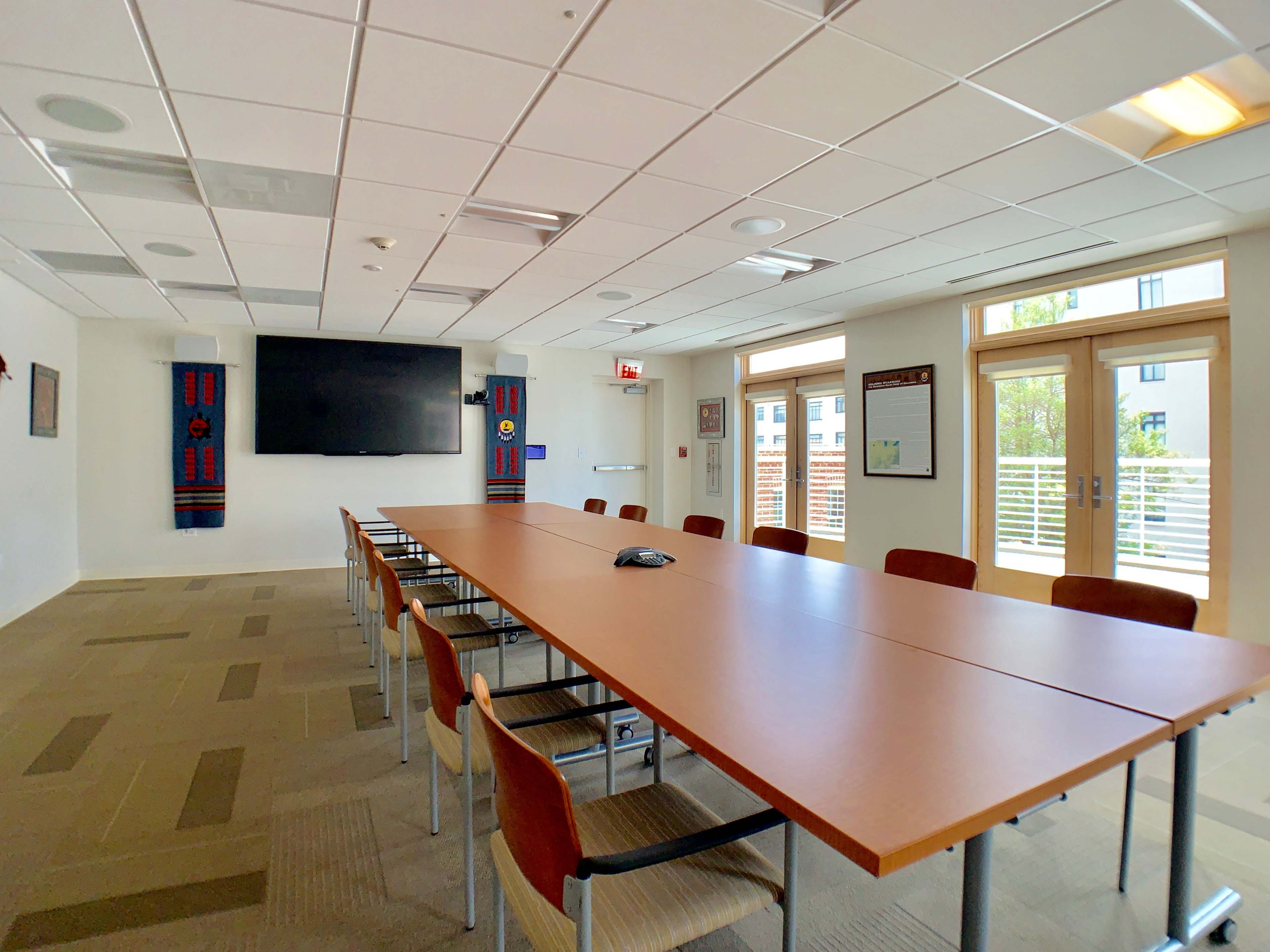A spacious conference room features a long wooden table, chairs, a large wall-mounted screen, and windows that open to an outdoor view.