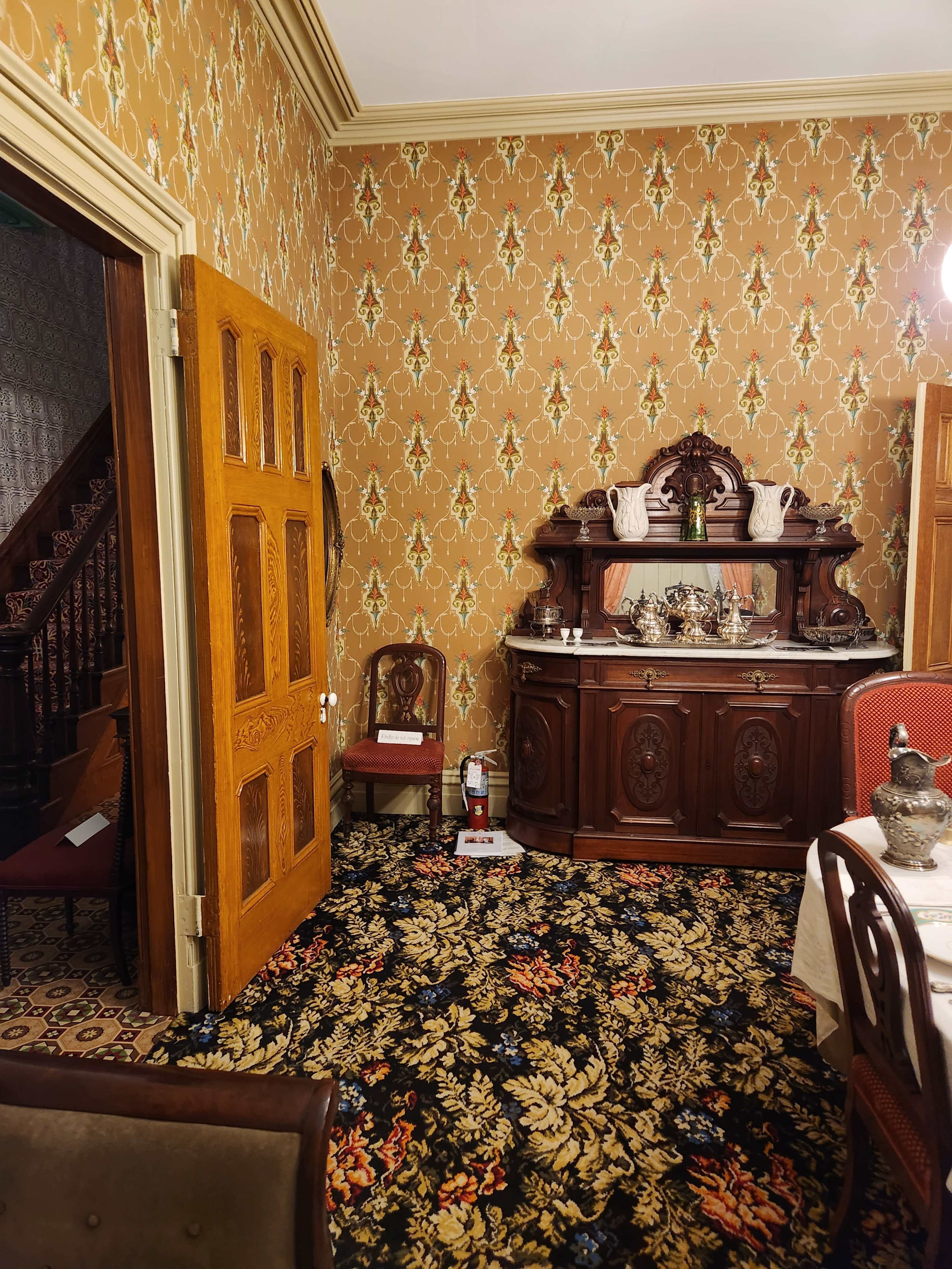A vintage dining room with ornate wallpaper, a wooden buffet, and a patterned carpet.