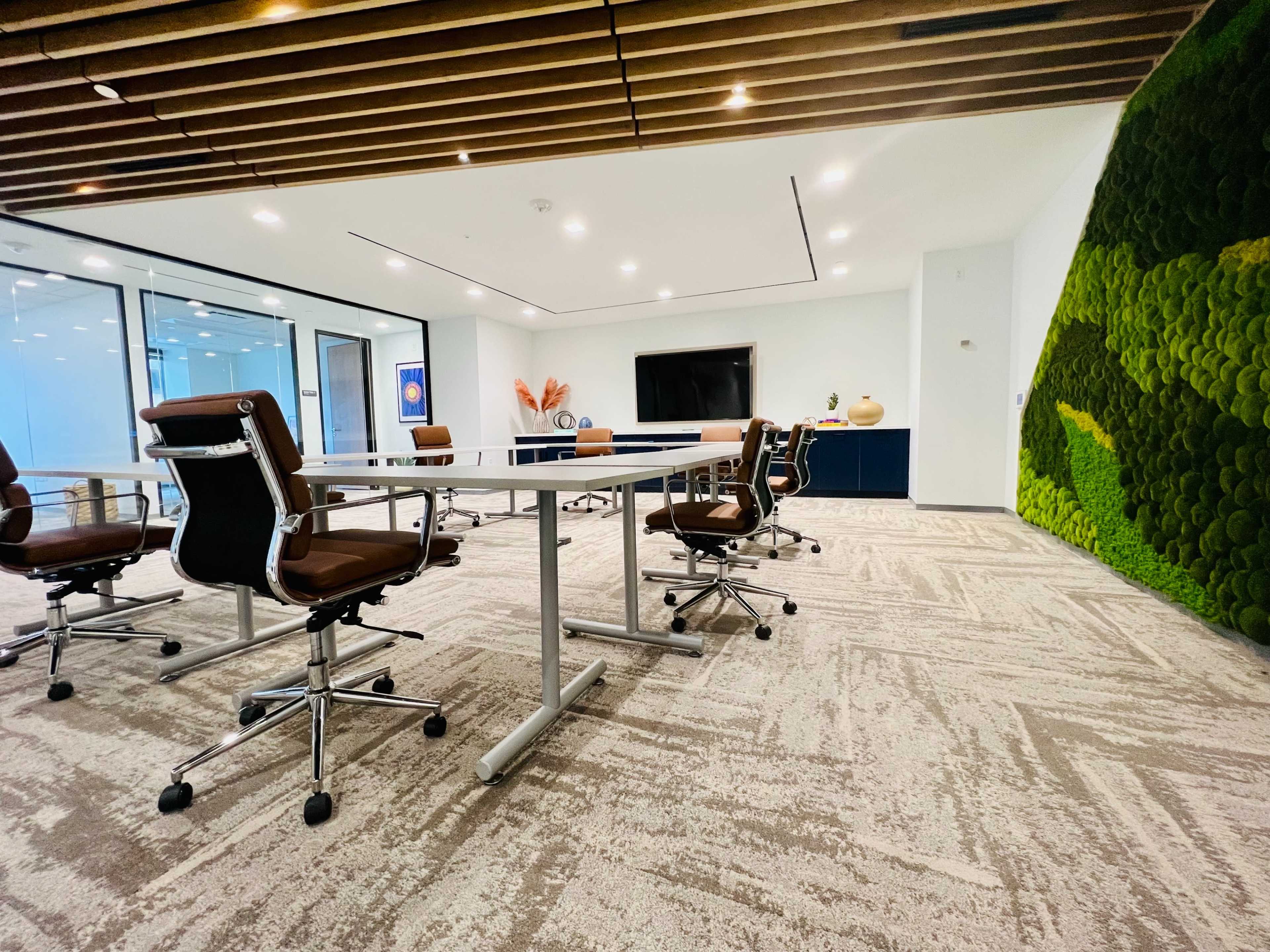 A modern conference room with a large table surrounded by rolling chairs, featuring a green moss wall and a television screen on one side.