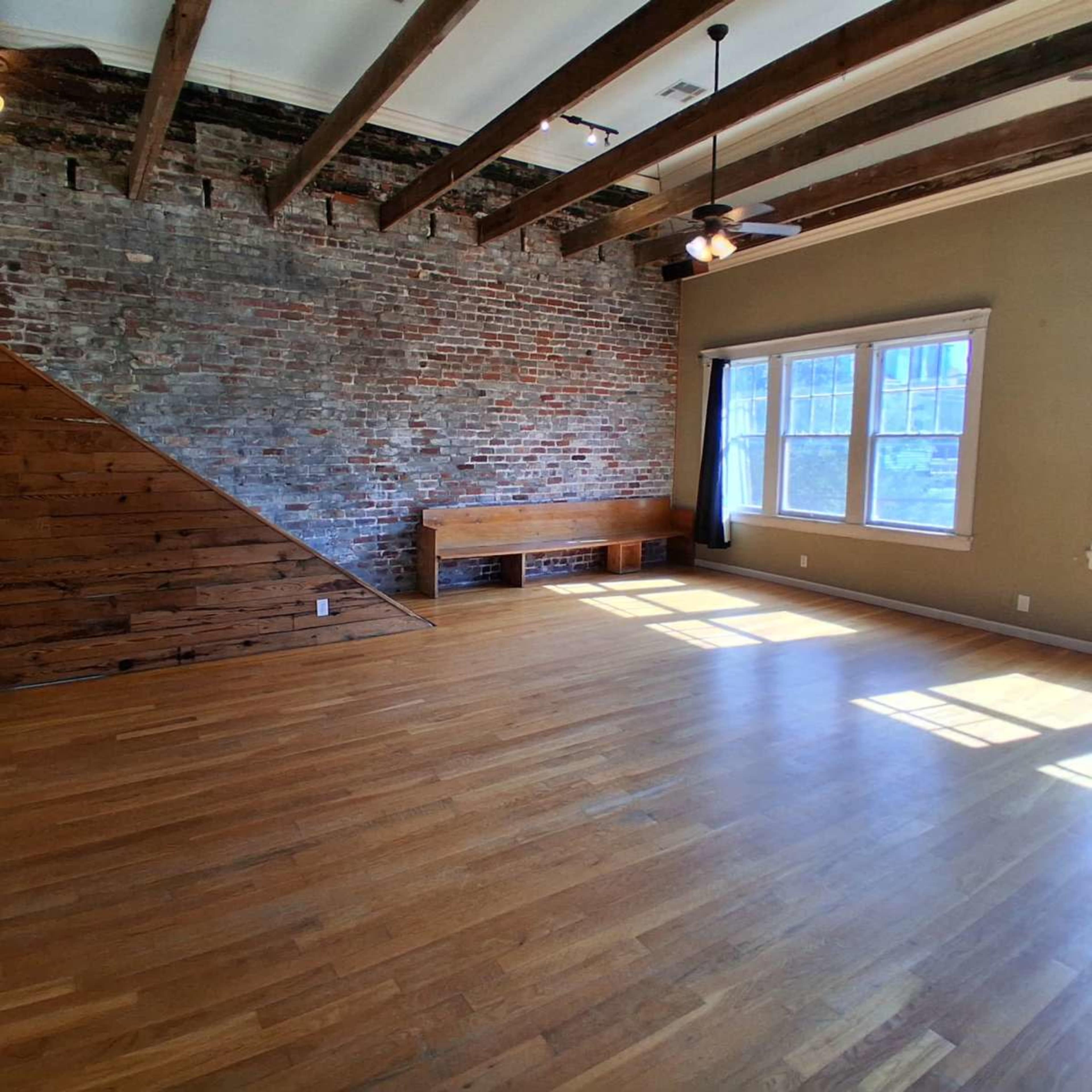 The image shows a spacious room with exposed brick walls, wooden beams, and large windows allowing natural light to flood in, along with a wooden bench against one wall.