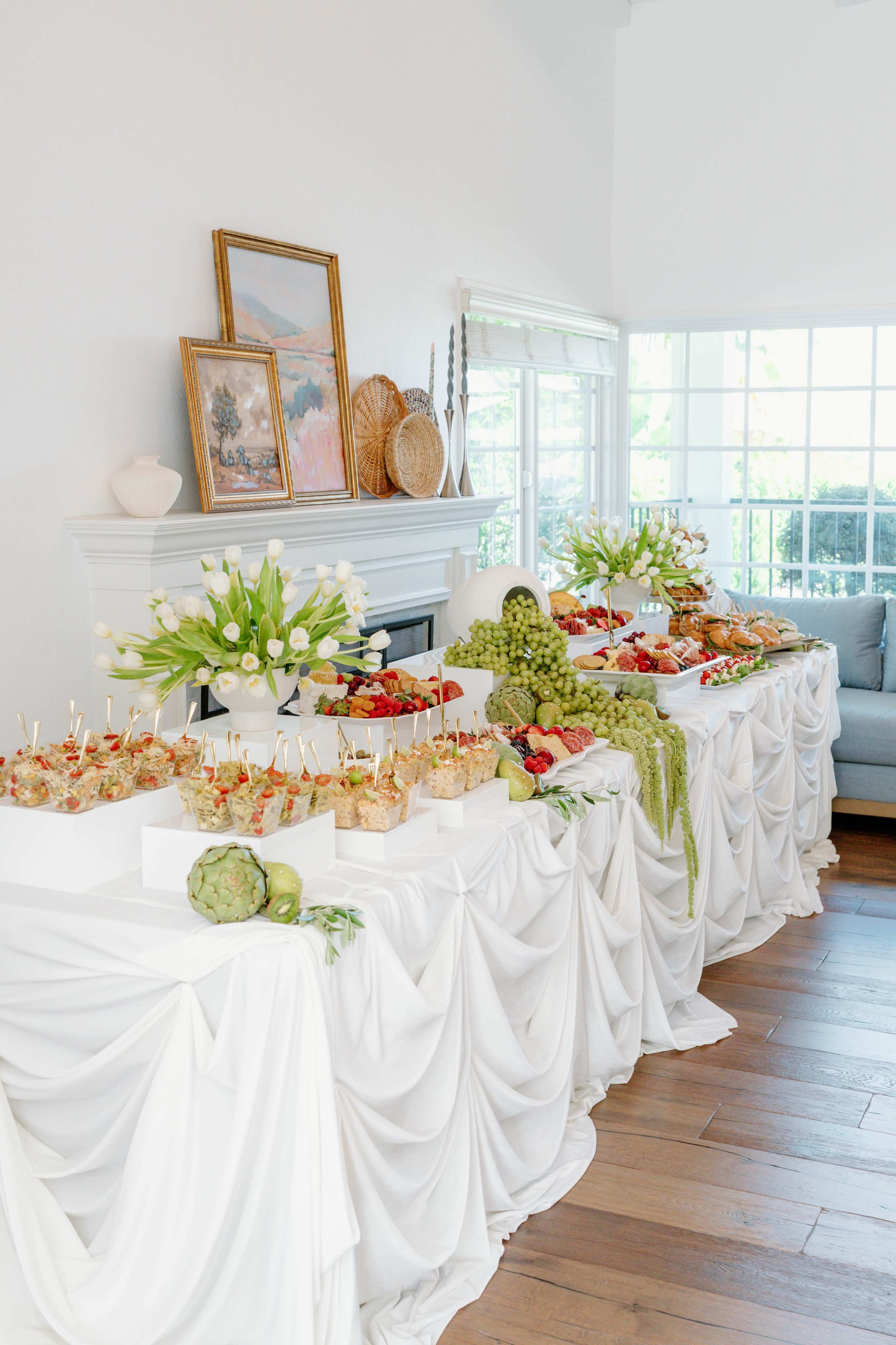 The image shows a beautifully arranged buffet table with an assortment of food, including fruits, vegetables, and gourmet appetizers, draped in white tablecloths.