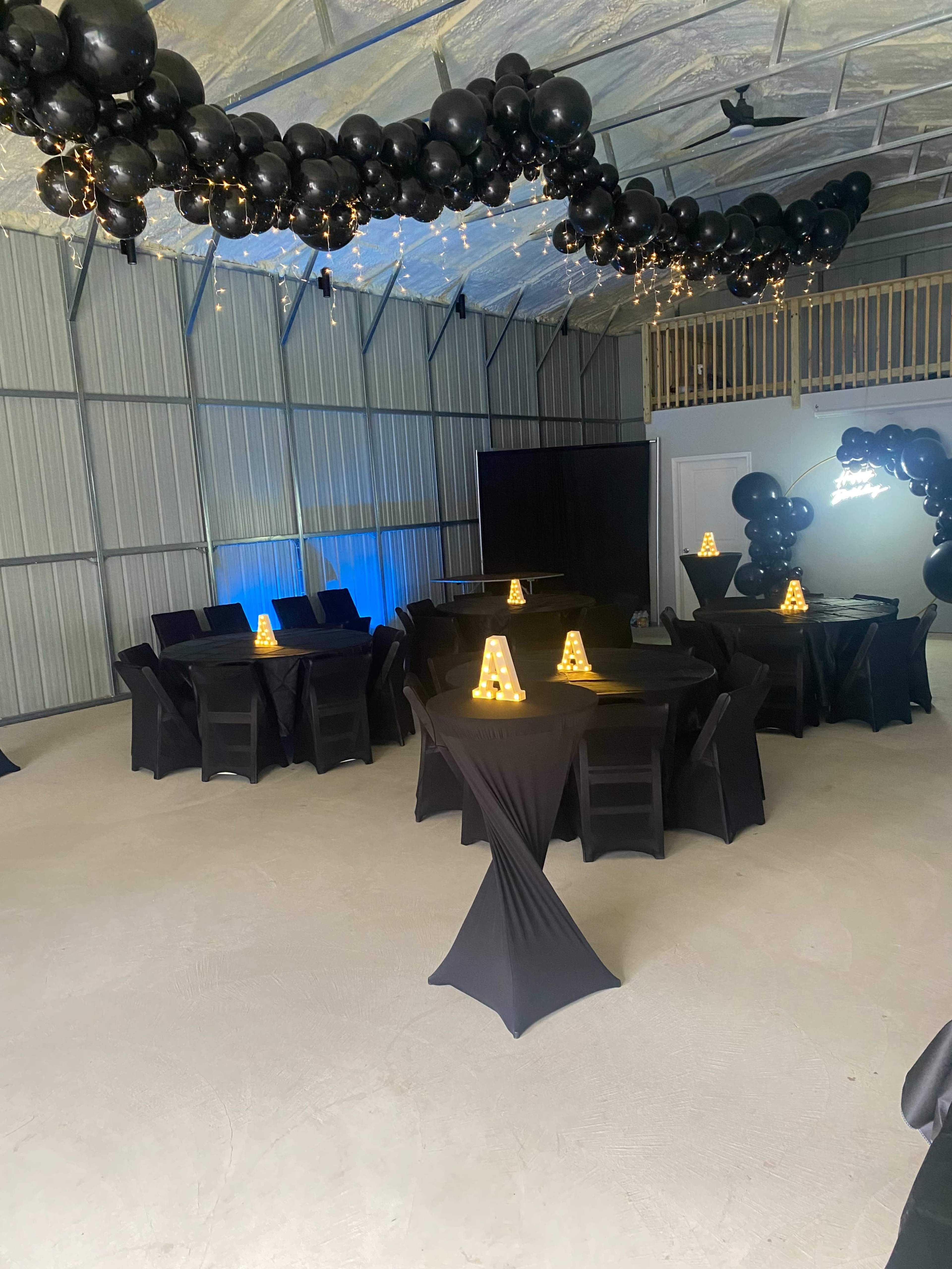 The image shows a spacious indoor venue with several black tables, each adorned with illuminated centerpieces, and a decorative display of black balloons and string lights overhead.