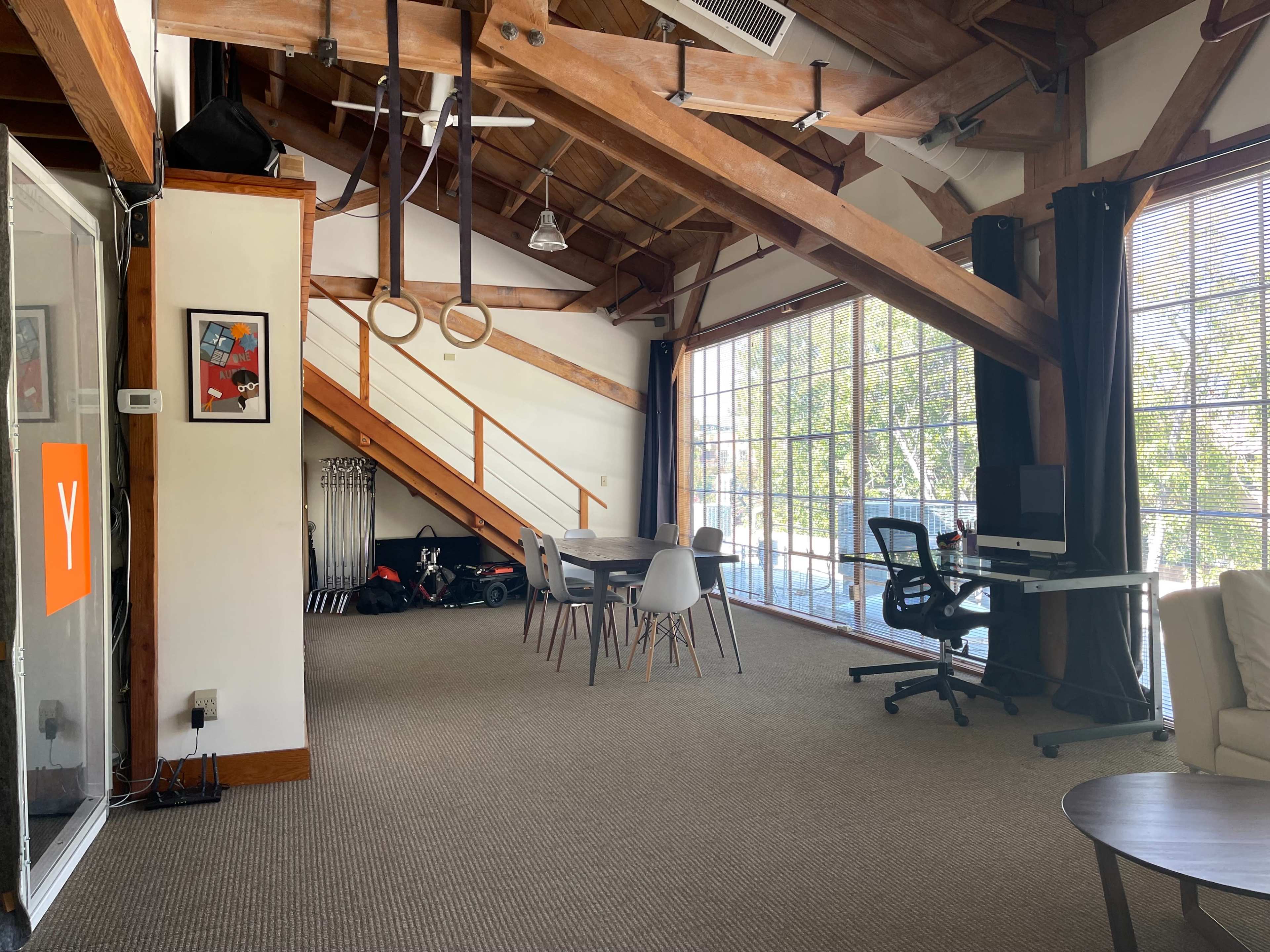 An open office space with wooden beams, large windows, a meeting table, and exercise rings hanging from the ceiling.