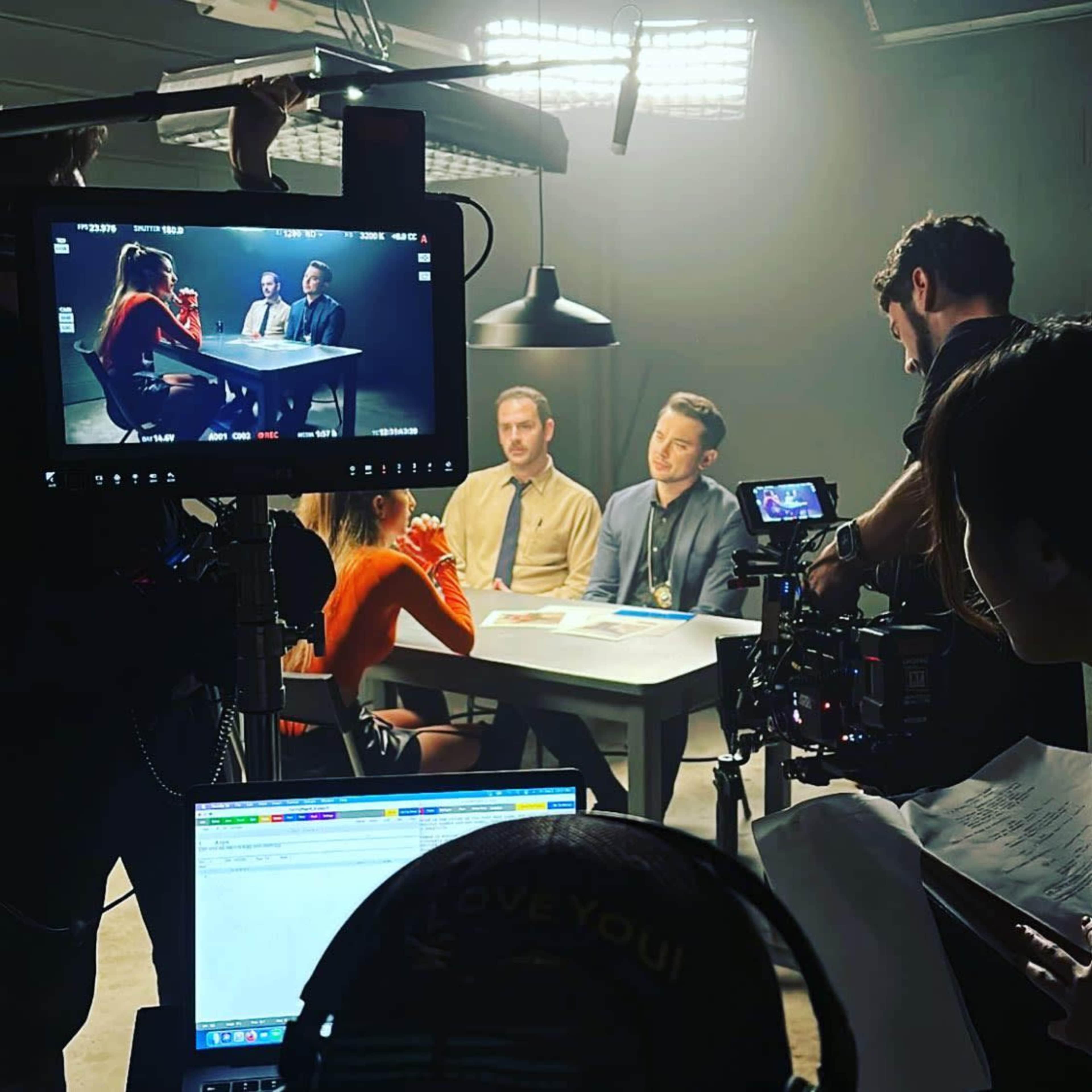 A film crew records a scene featuring three actors sitting at a table under studio lighting.