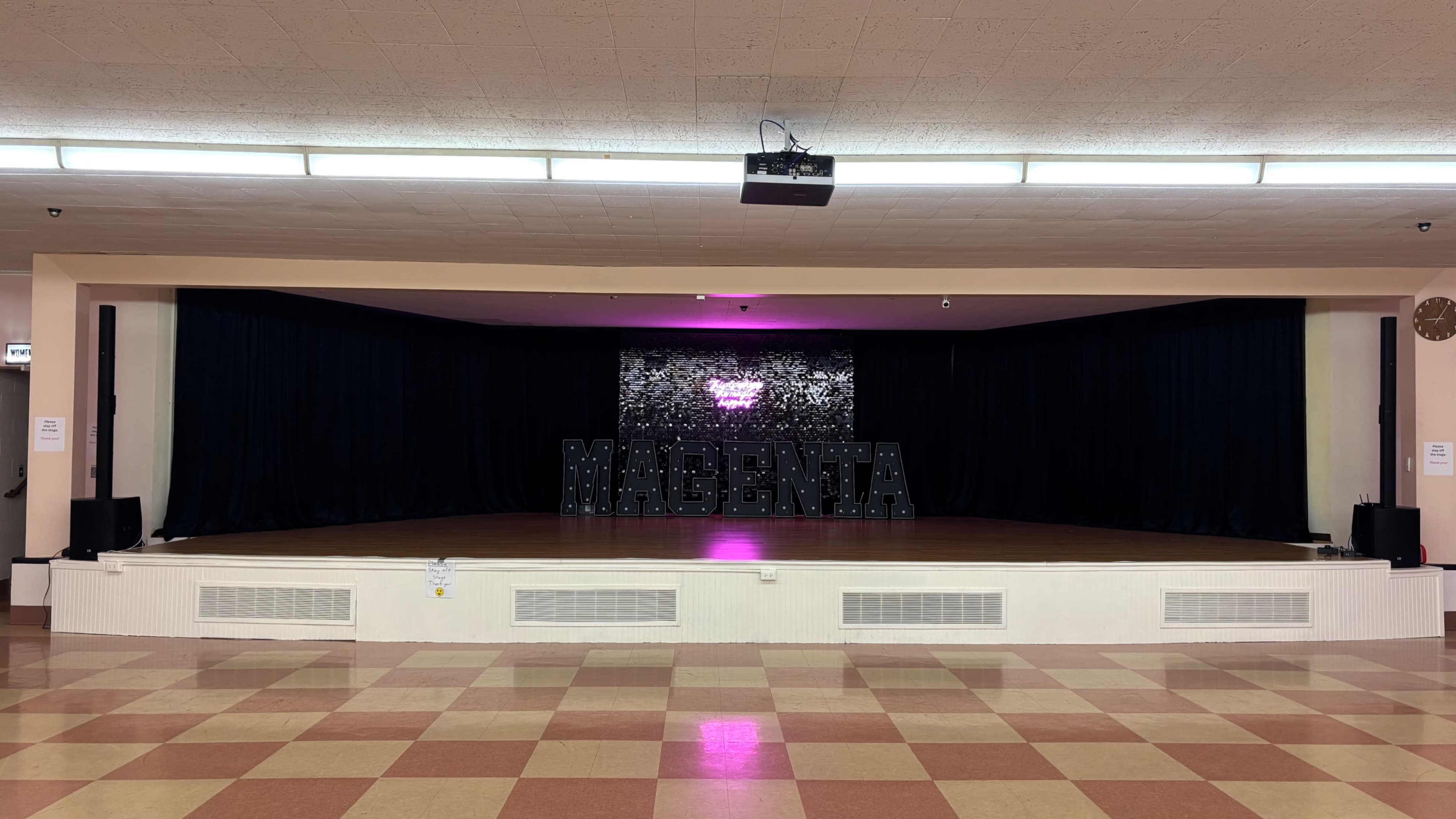 The image shows a stage with a black backdrop, a large decorative "MAGENTA" sign, and a sound system on either side, set in a hall with a checkered floor.