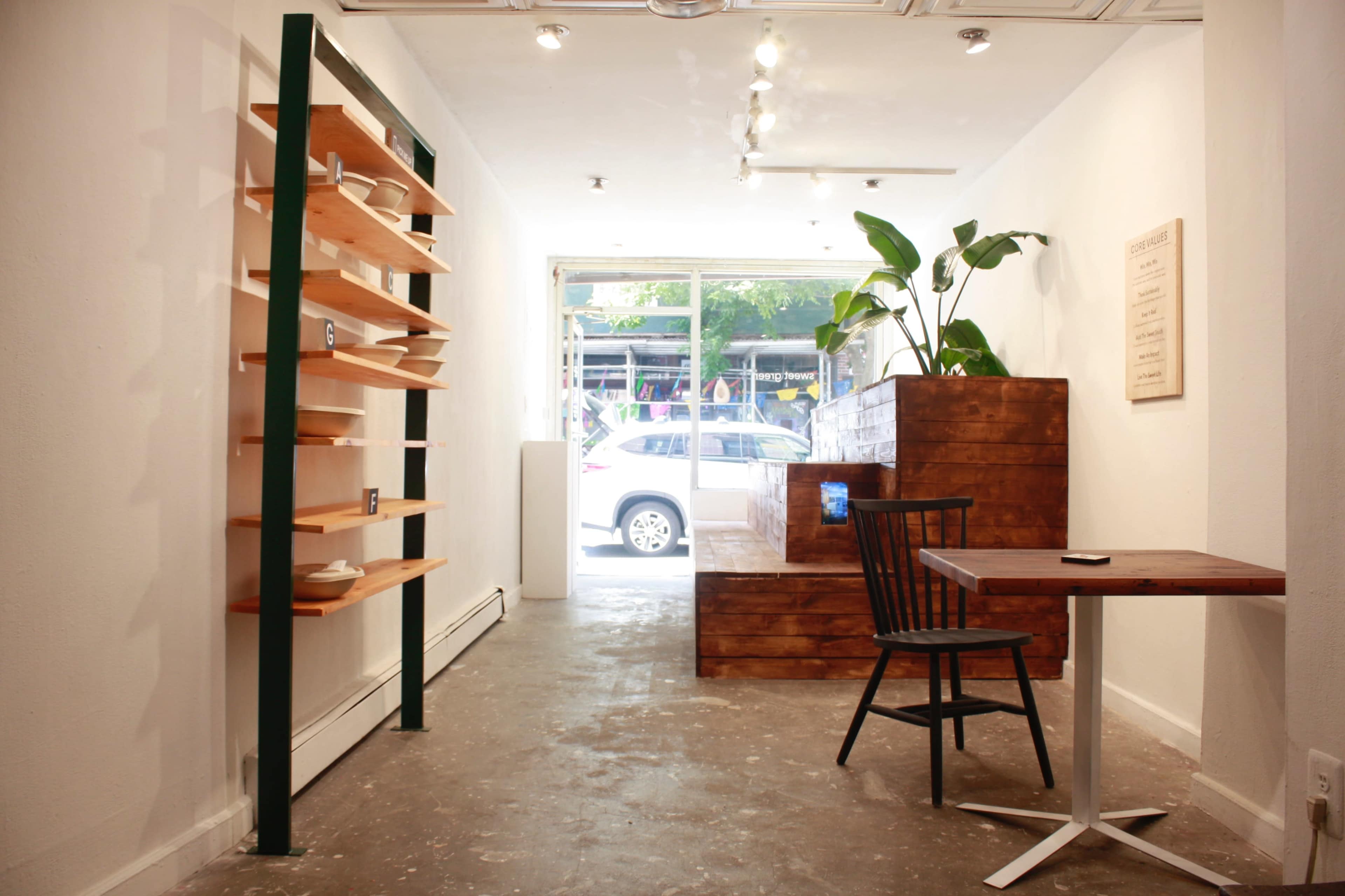 A minimalist interior of a café or shop featuring wooden shelving, a wooden seating area, and a small table with a chair, along with large windows displaying a street view.
