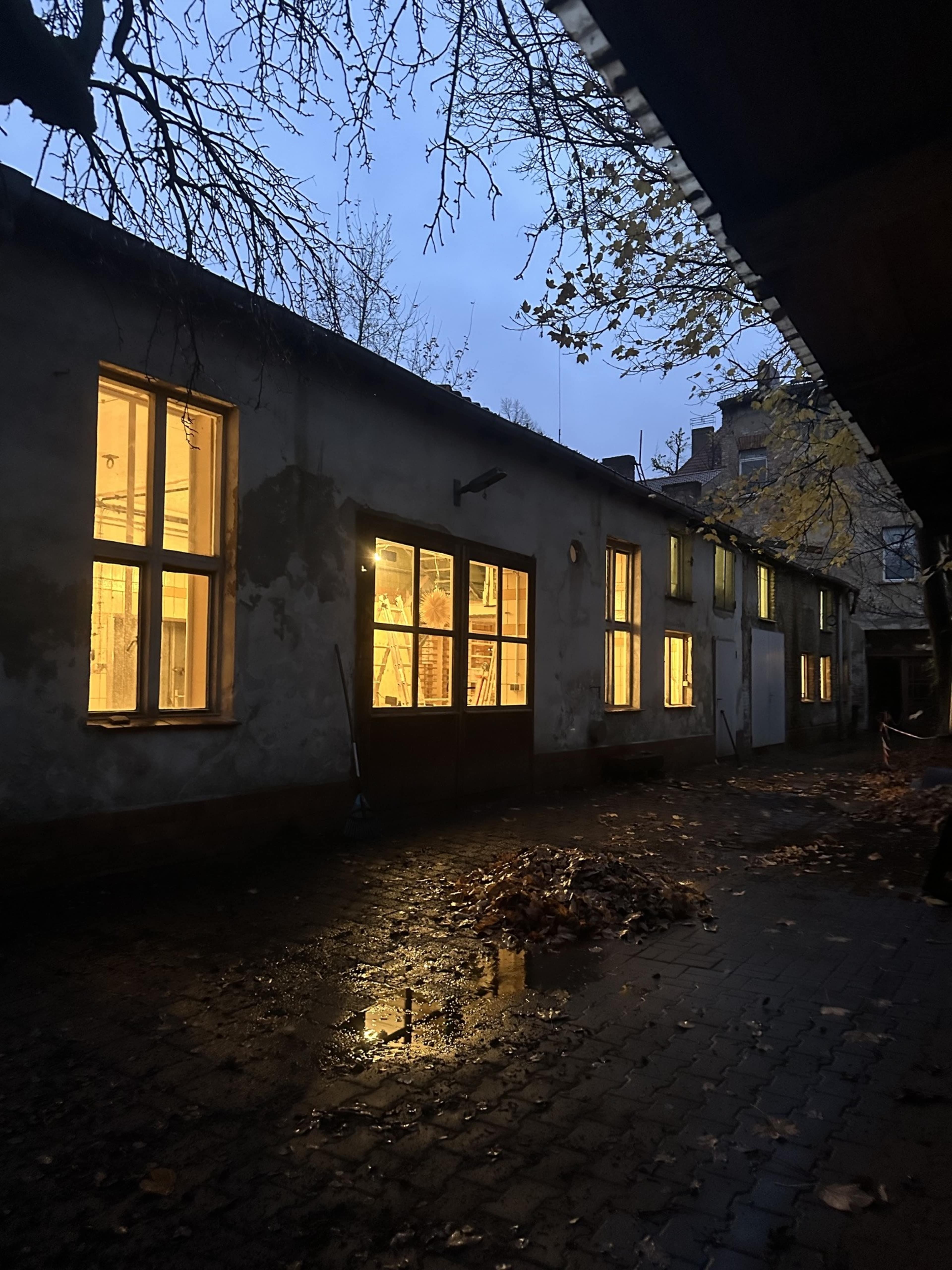 The image shows a dimly lit building with several illuminated windows reflecting on a wet cobblestone path, surrounded by autumn leaves.