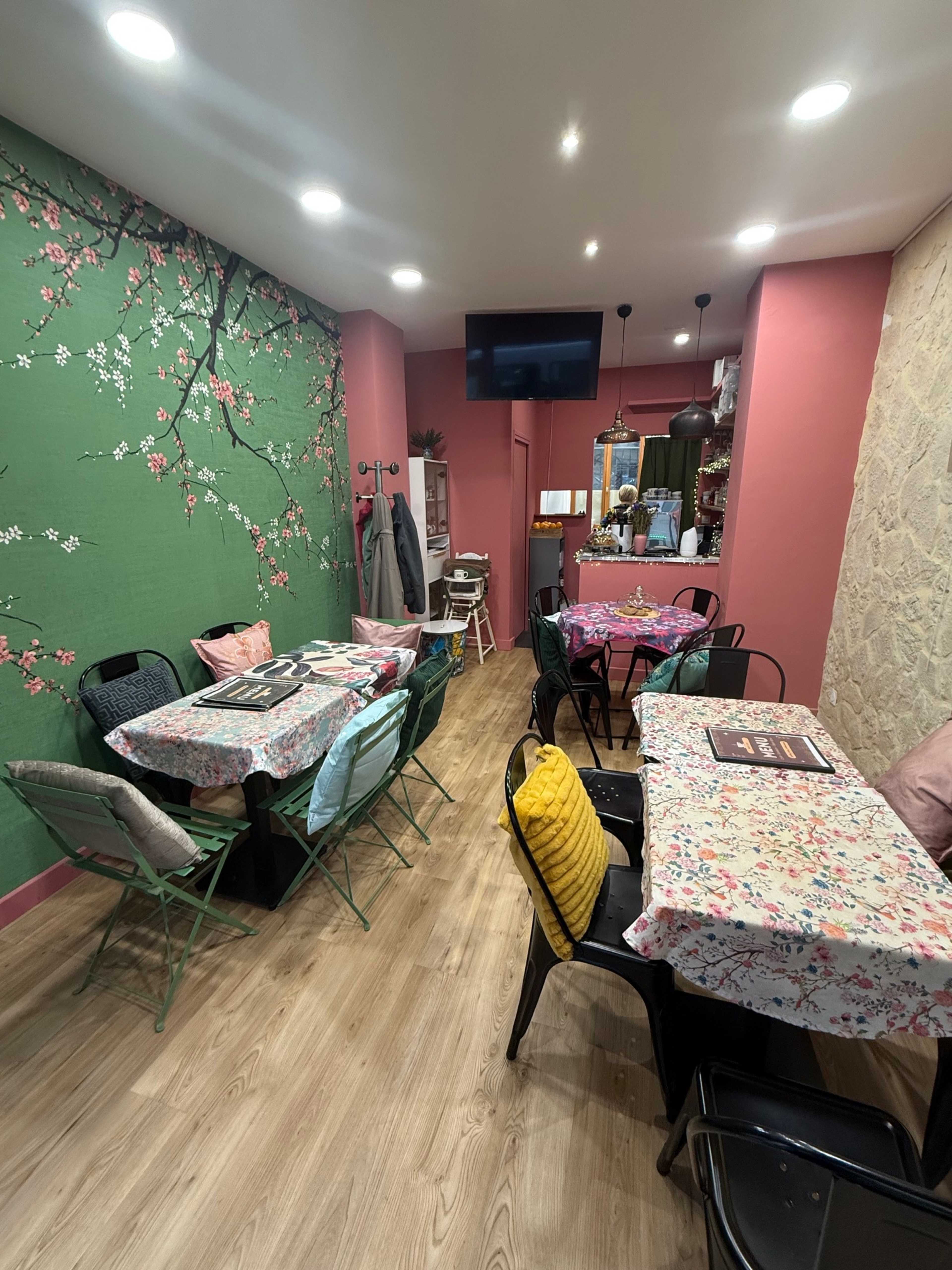 The image shows a small, cozy restaurant with floral-patterned tablecloths, colorful chairs, and green walls adorned with a mural of cherry blossoms.