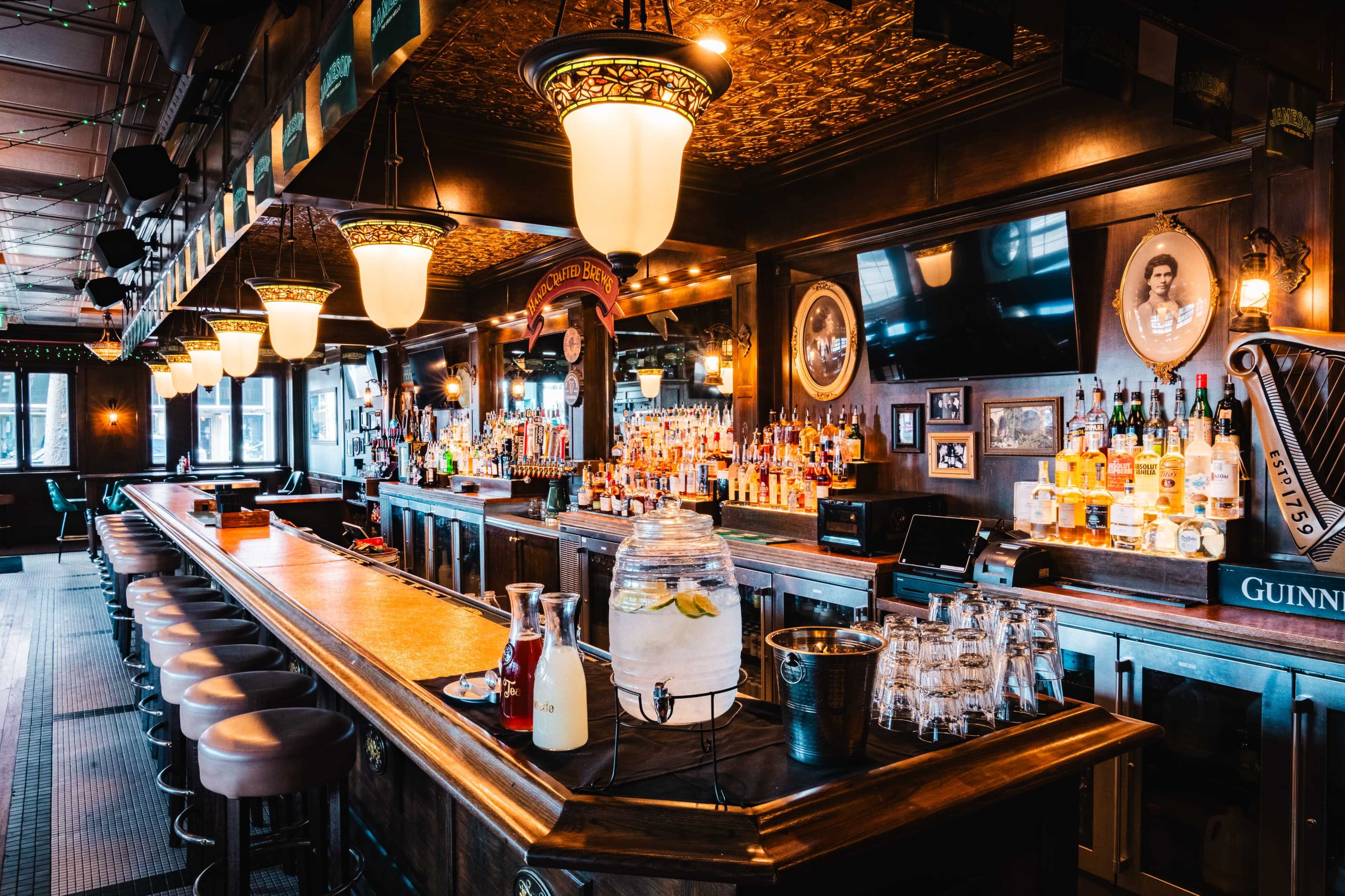 A wooden bar with a variety of liquor bottles on shelves, illuminated by overhead lights, features a large counter and several bar stools.