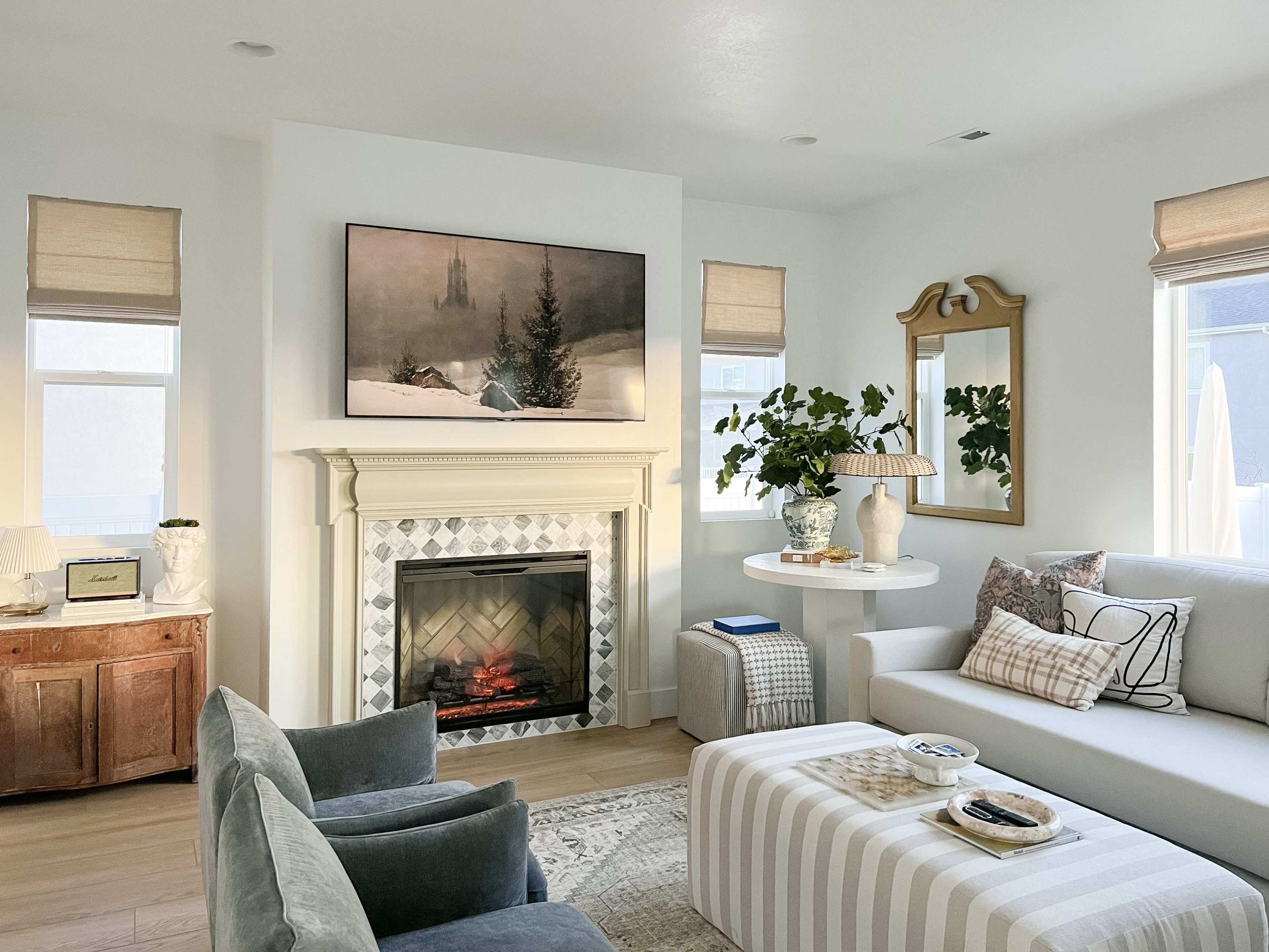A modern living room featuring a fireplace with a TV mounted above it, a round table with a vase of greenery, a sofa with decorative pillows, and a wooden cabinet.