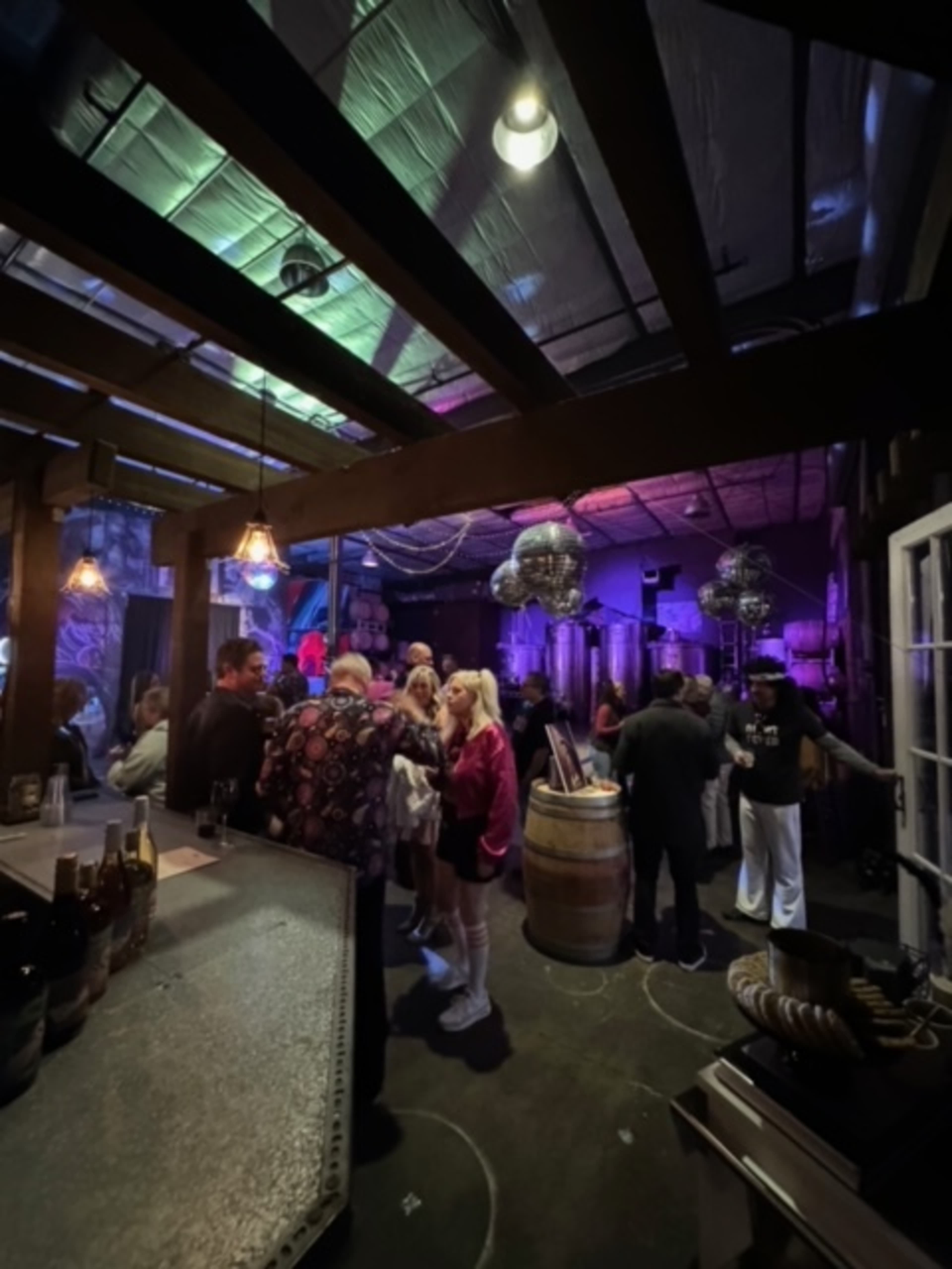 A group of people gathers in a dimly lit bar with colorful lights and disco balls, surrounded by barrels and a rustic wooden structure.