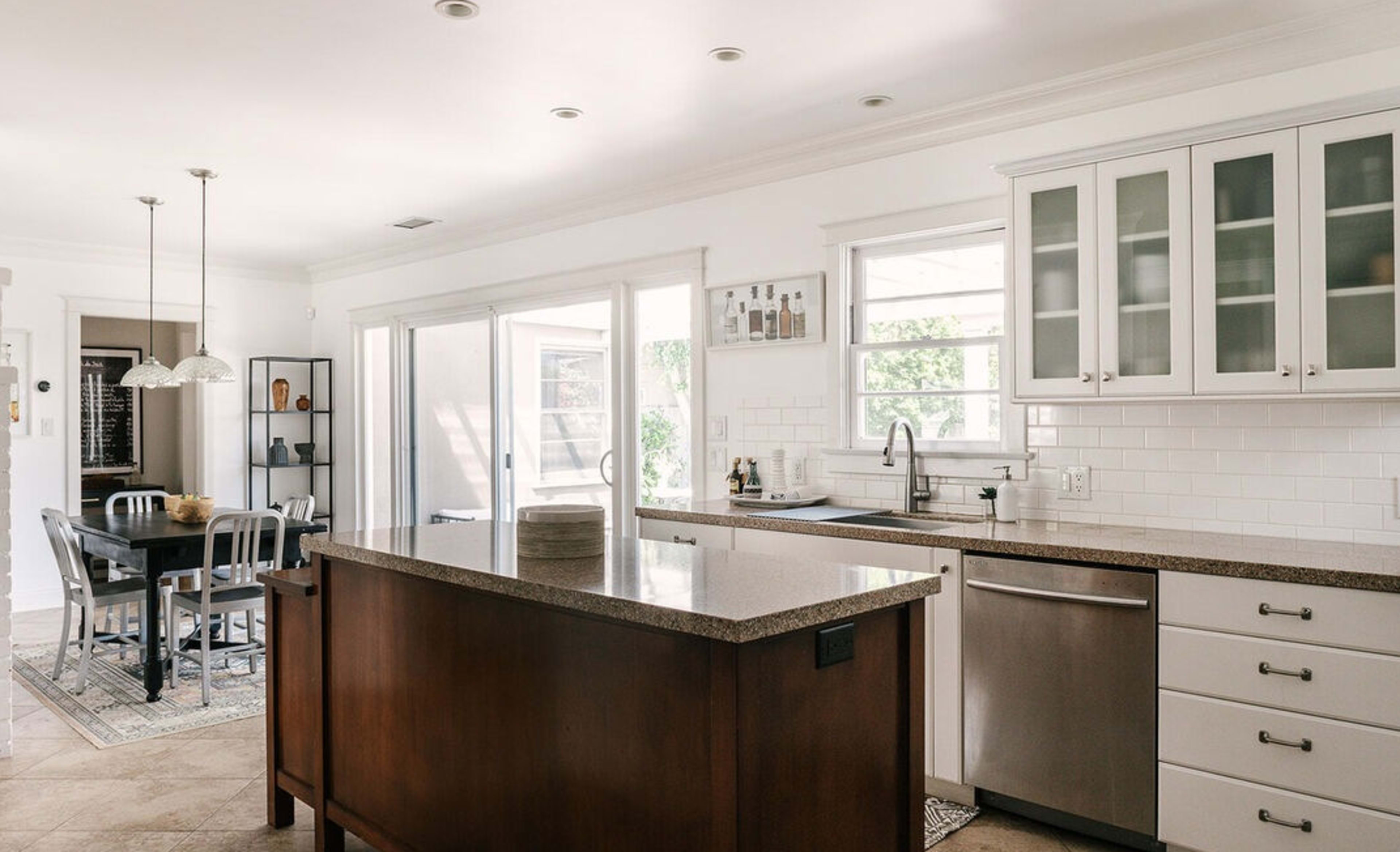 A modern kitchen features a central island with a granite countertop, stainless steel appliances, and white cabinetry, leading to a dining area with a wooden table and metal chairs.
