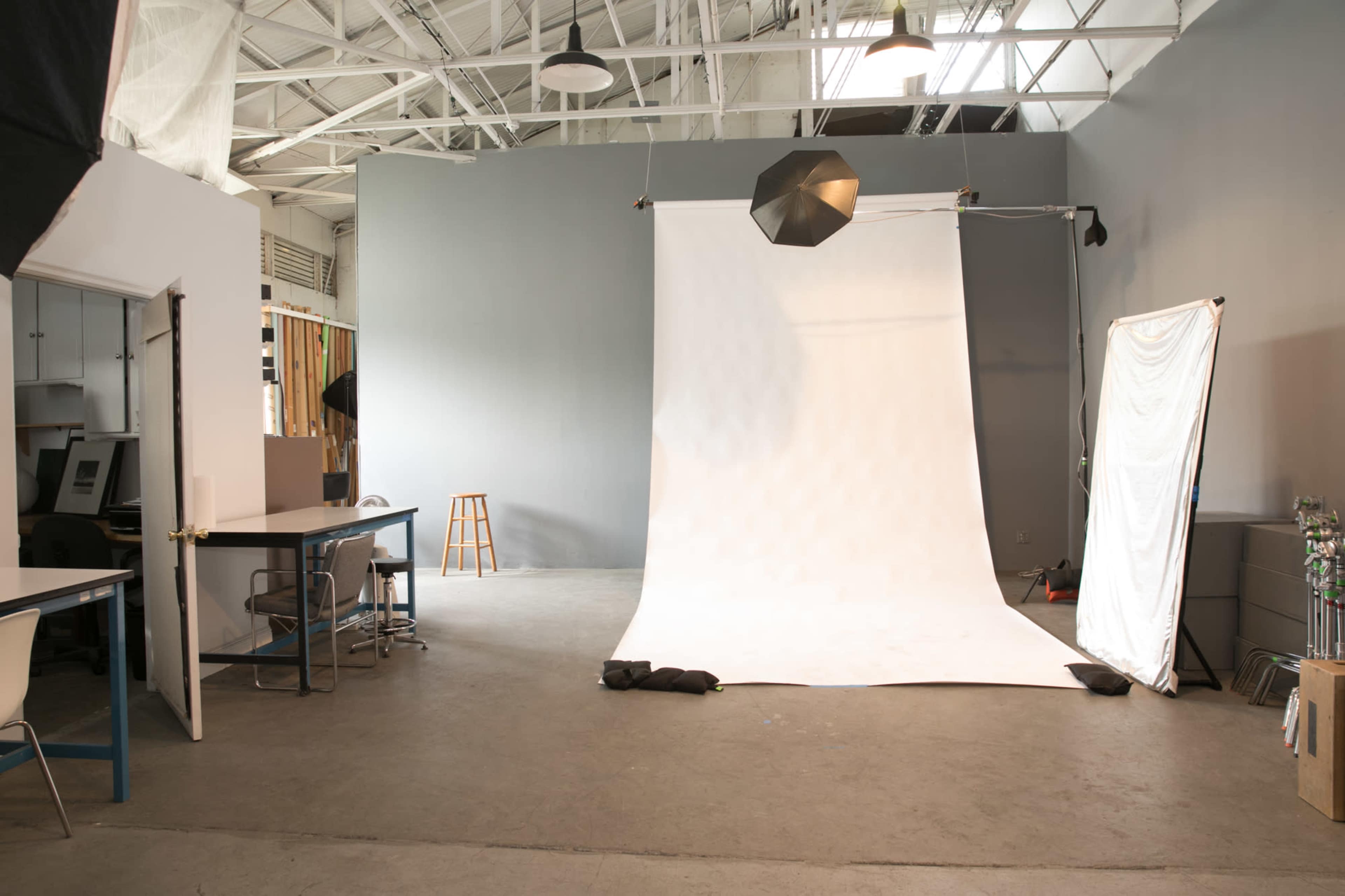 A photography studio features a large white backdrop, a stool, studio lights, and a workspace with tables and chairs.