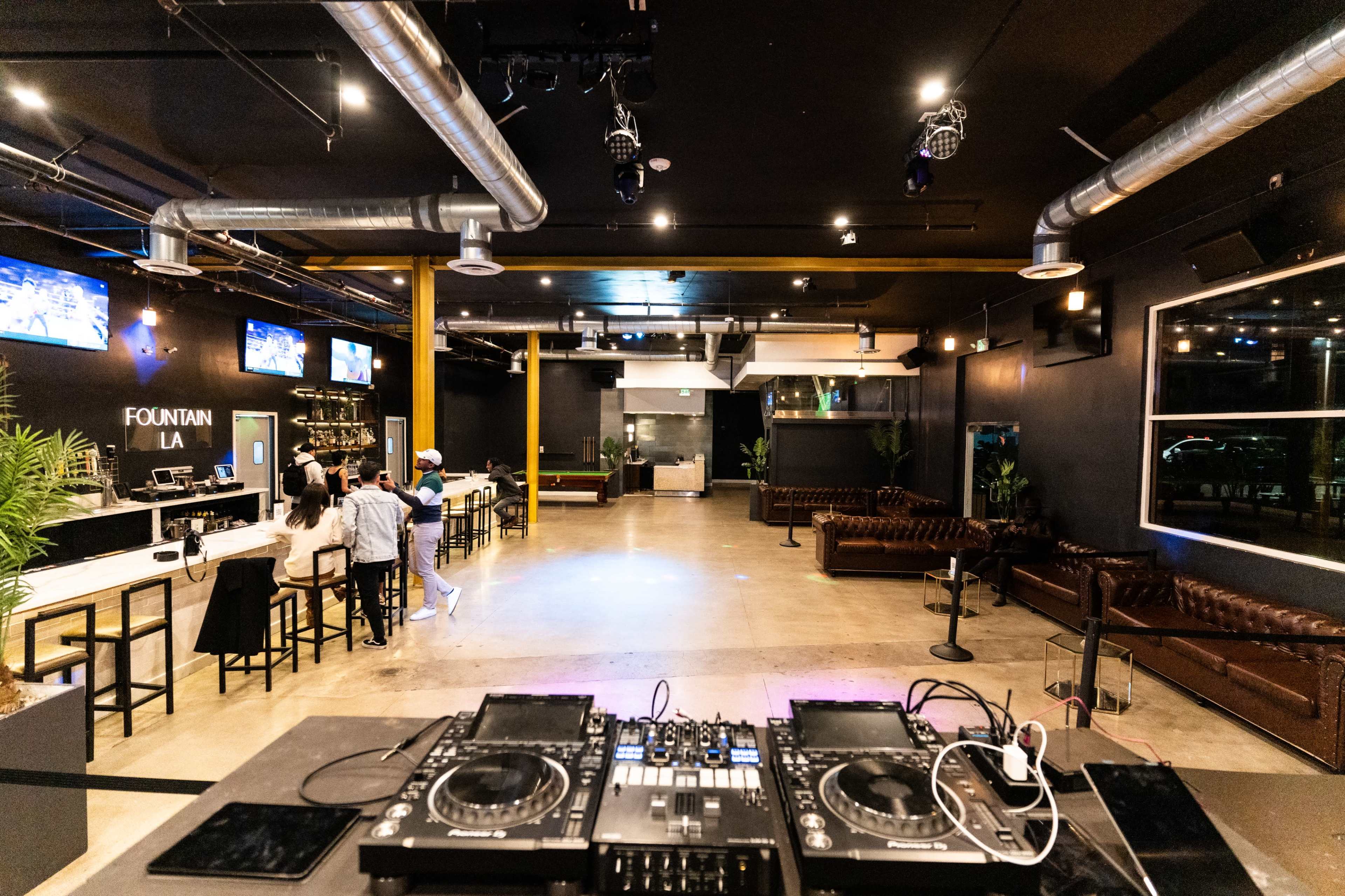 A DJ booth in a dimly lit venue with seating areas, a bar, and large screens on the walls.