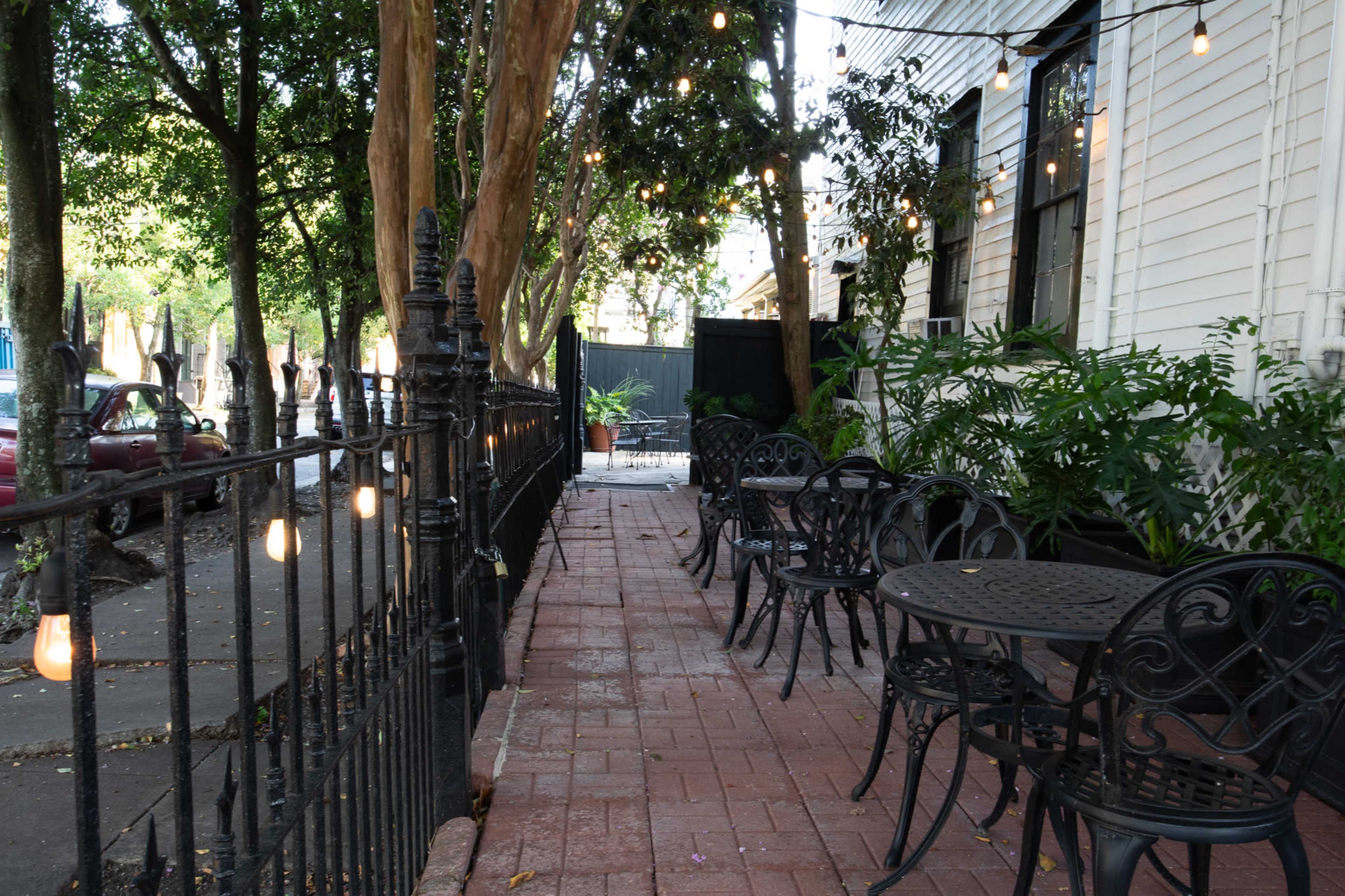 The image shows a brick pathway lined with potted plants and outdoor tables surrounded by decorative iron fencing and hanging lights.