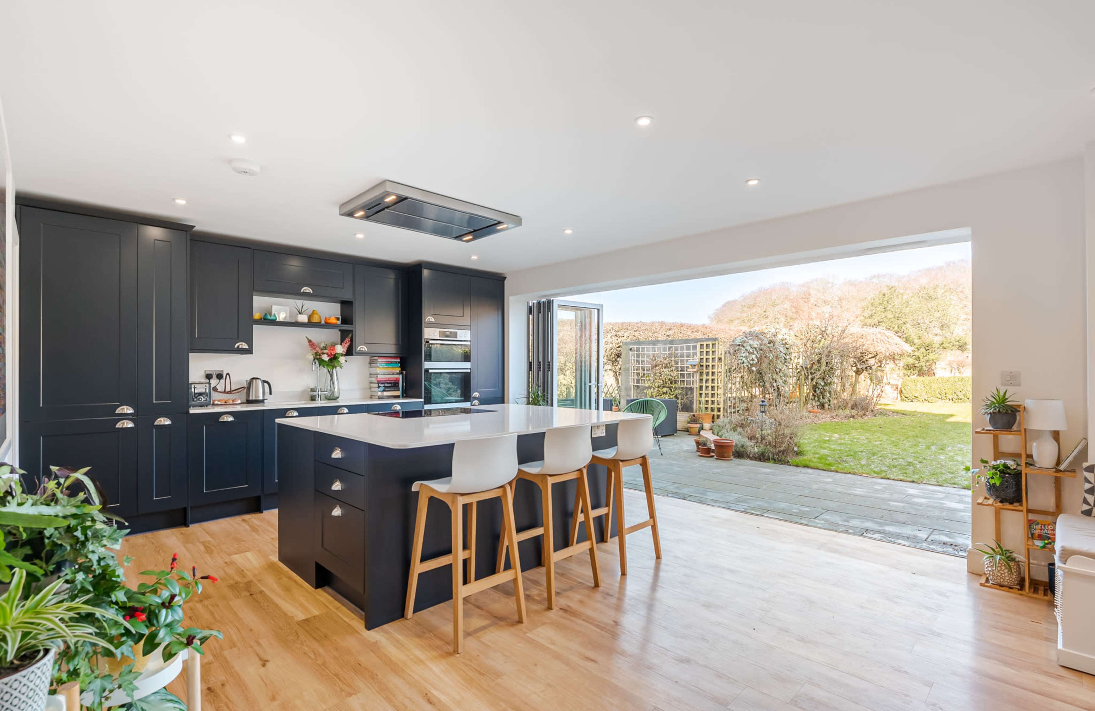 A modern kitchen features dark cabinetry, a central island with seating, and large glass doors that open to a garden.