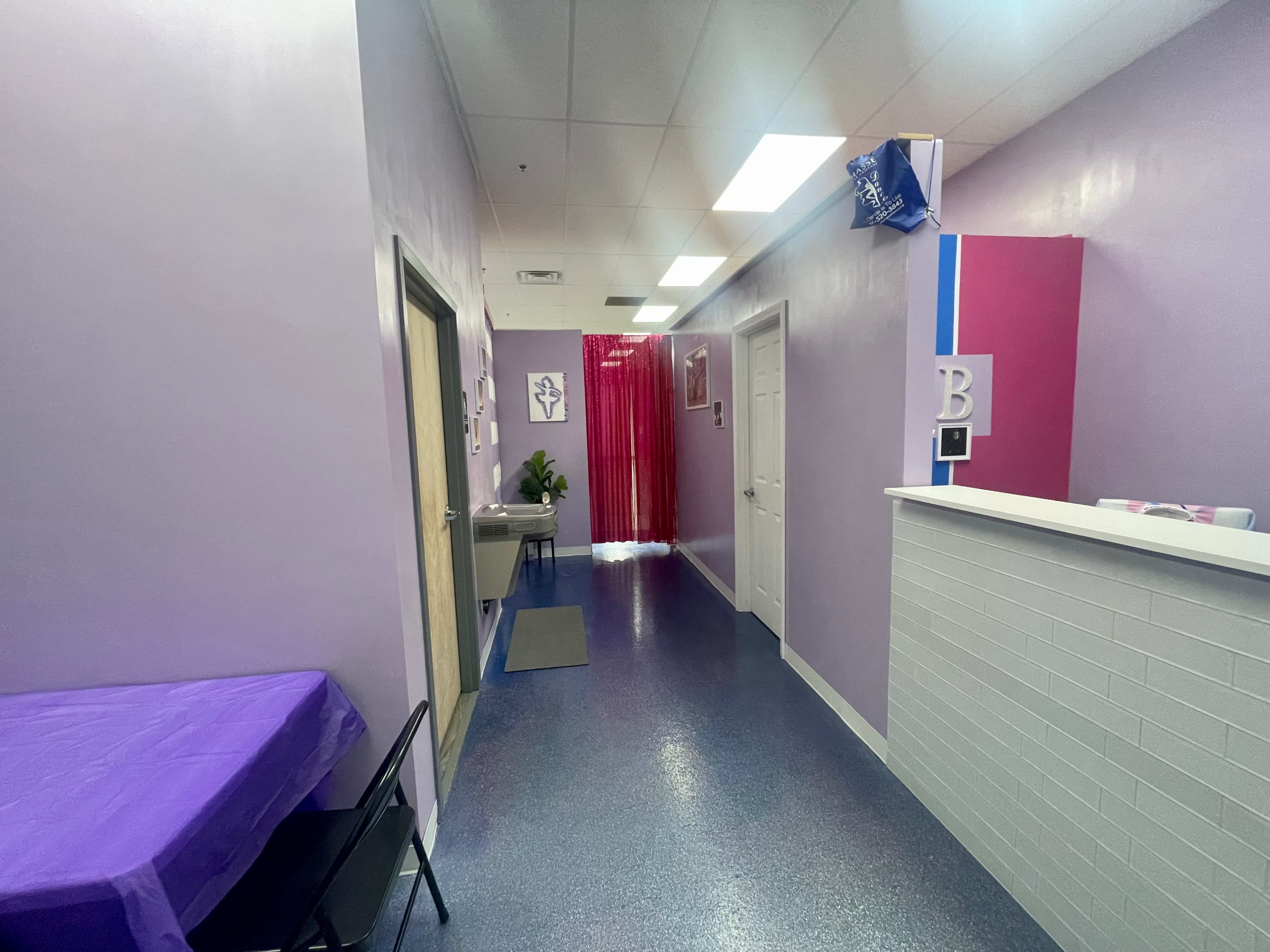 The image shows a brightly lit hallway in a salon or spa, featuring lavender walls, a reception desk at the end, and a purple table on the left side.