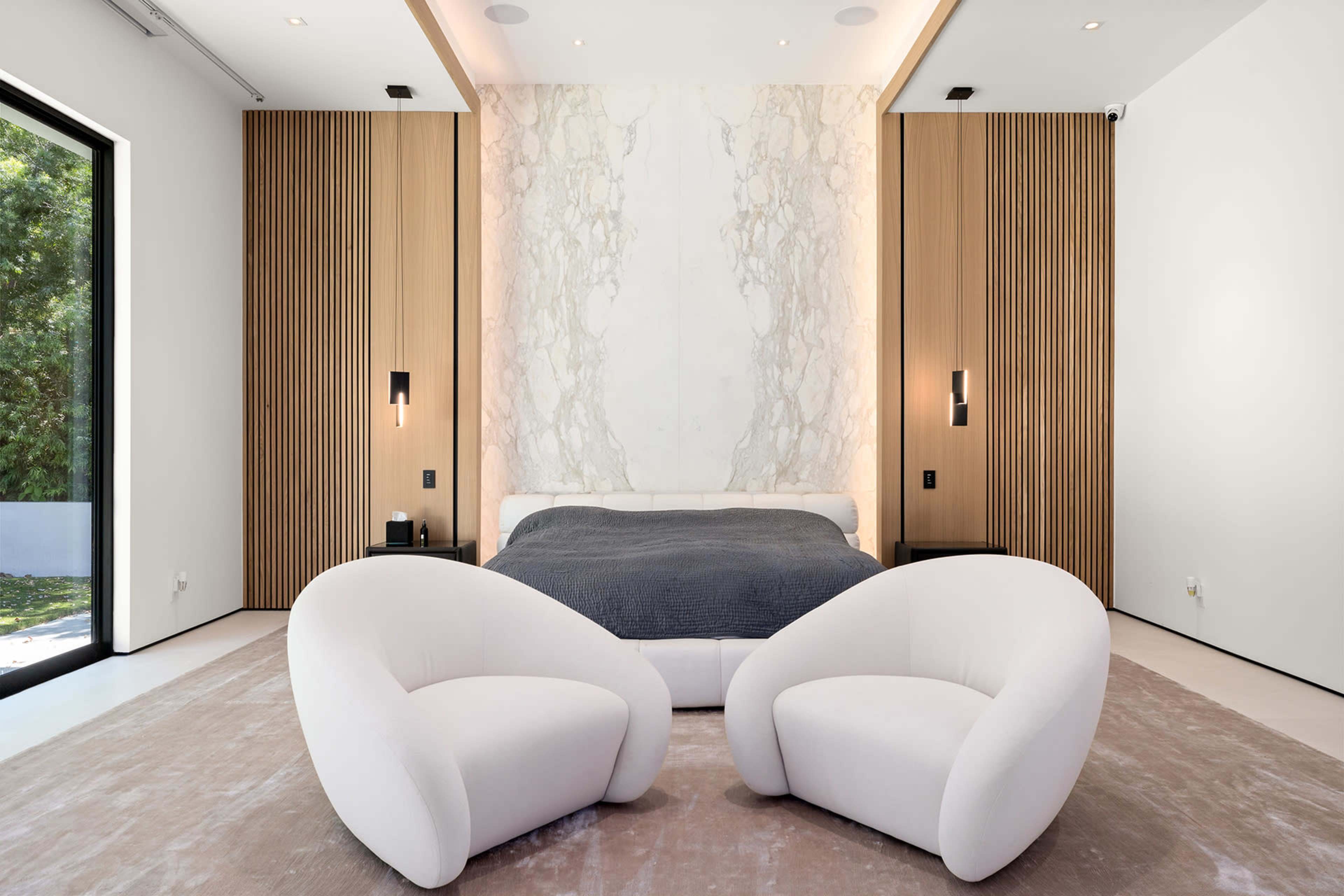 The image shows a modern bedroom featuring a bed against a marble accent wall, two curved chairs in front, and large windows letting in natural light.