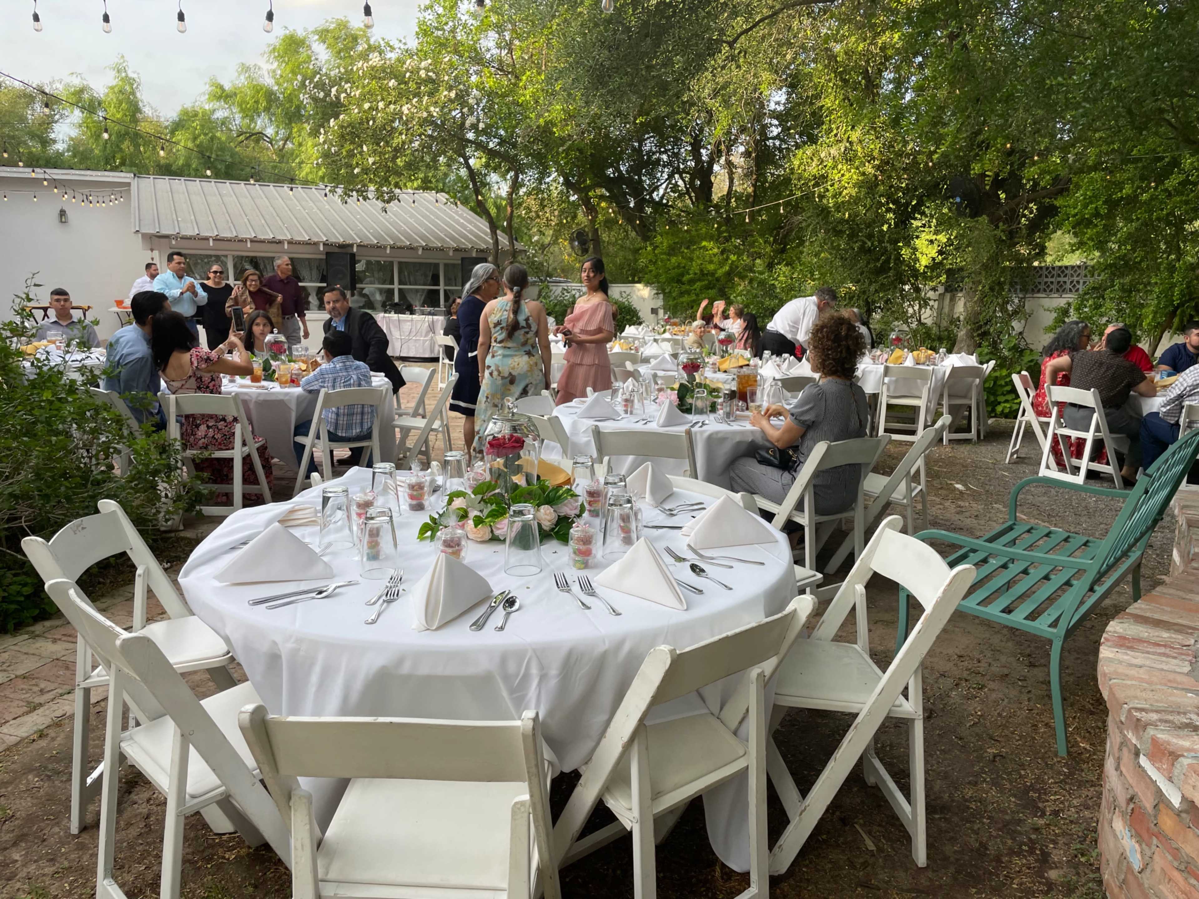 A garden event is taking place with guests seated at white-clothed tables surrounded by greenery and twinkling lights.
