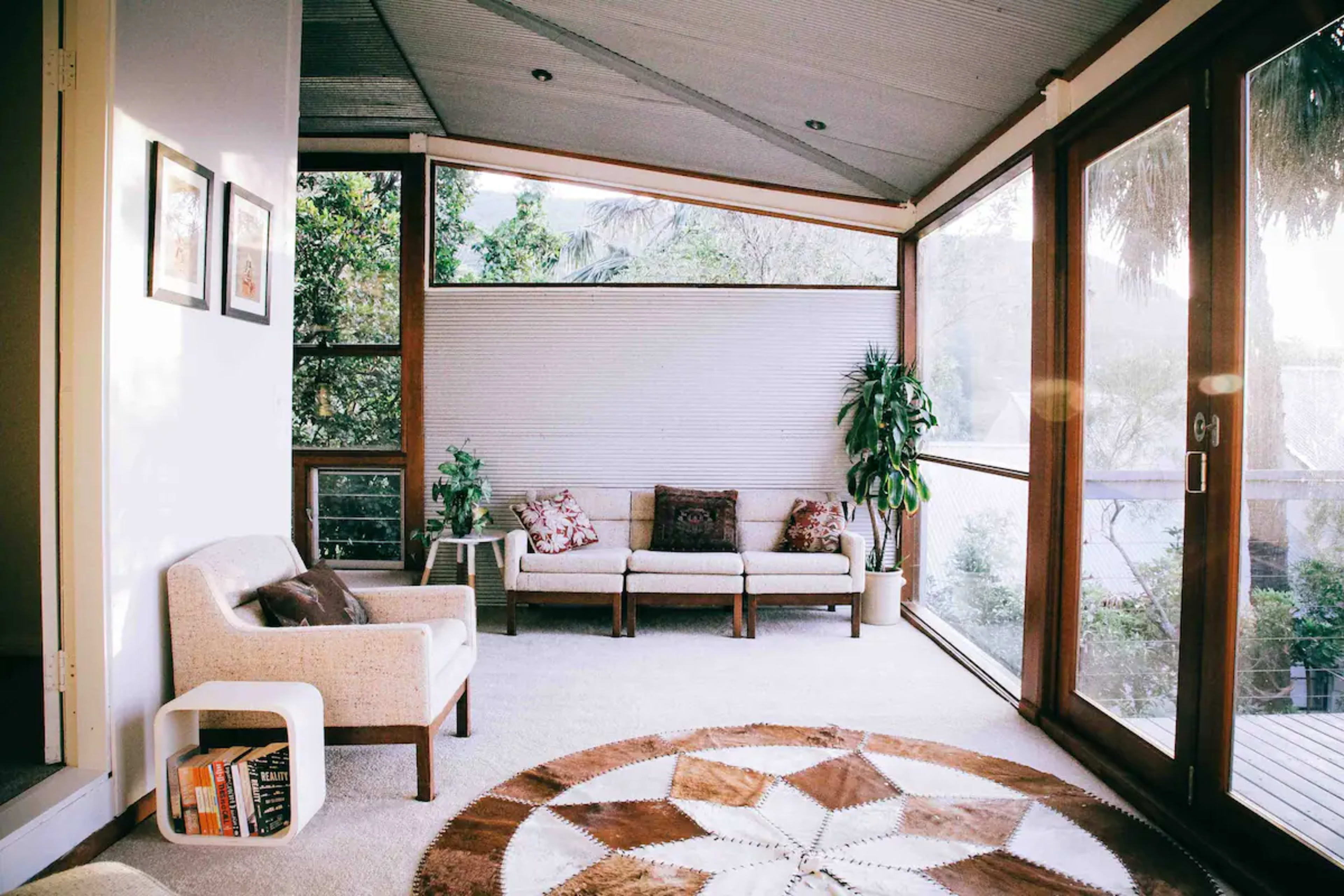A sunlit sitting area with a large window, featuring a couple of sofas, a plant, and a circular rug on the floor.