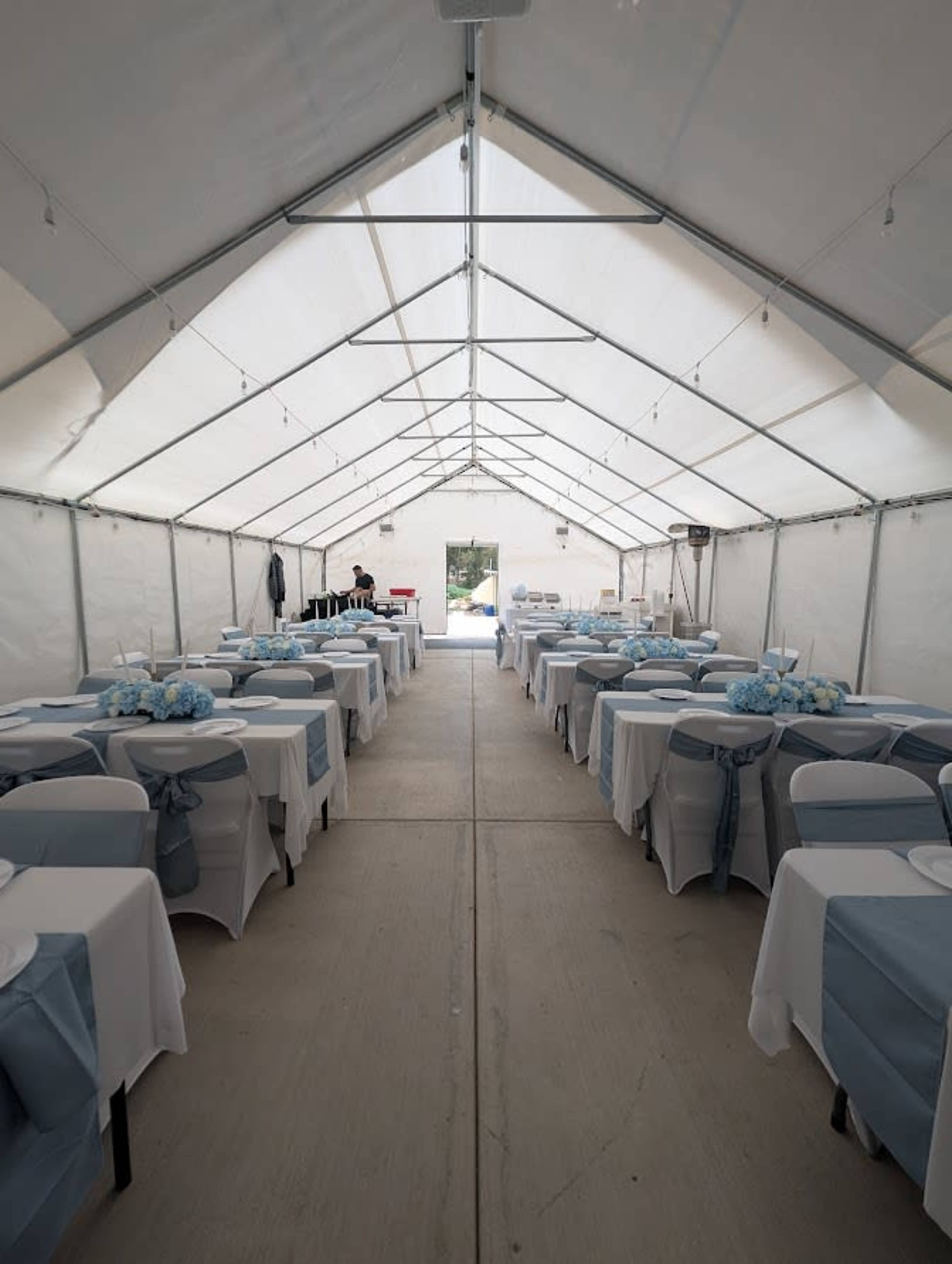 The image shows a tent set up for an event, with neatly arranged tables covered in white linens and blue sashes along the aisles.