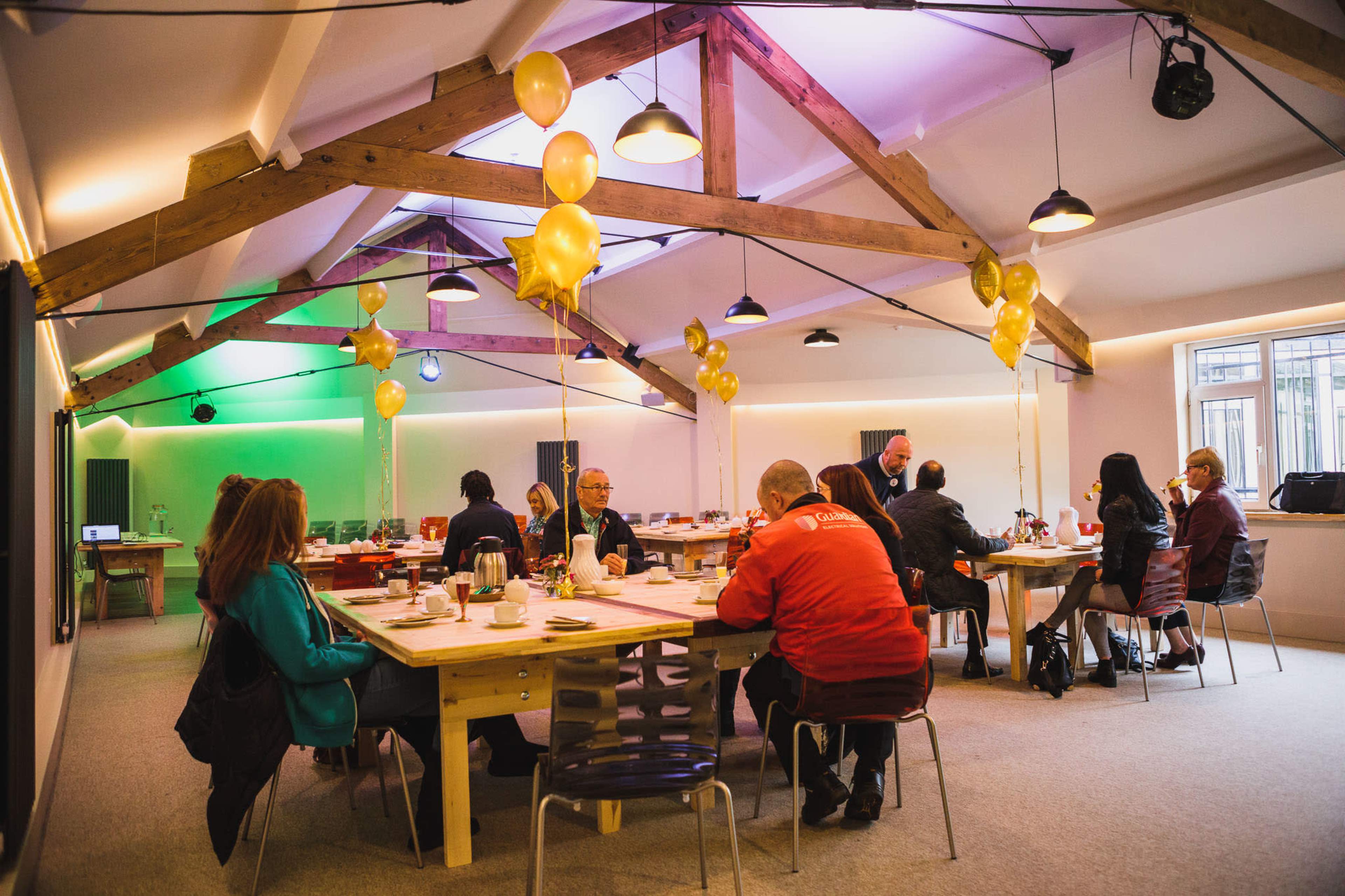 A bright event space features tables set for a gathering, decorated with yellow balloons and various attendees seated and mingling.
