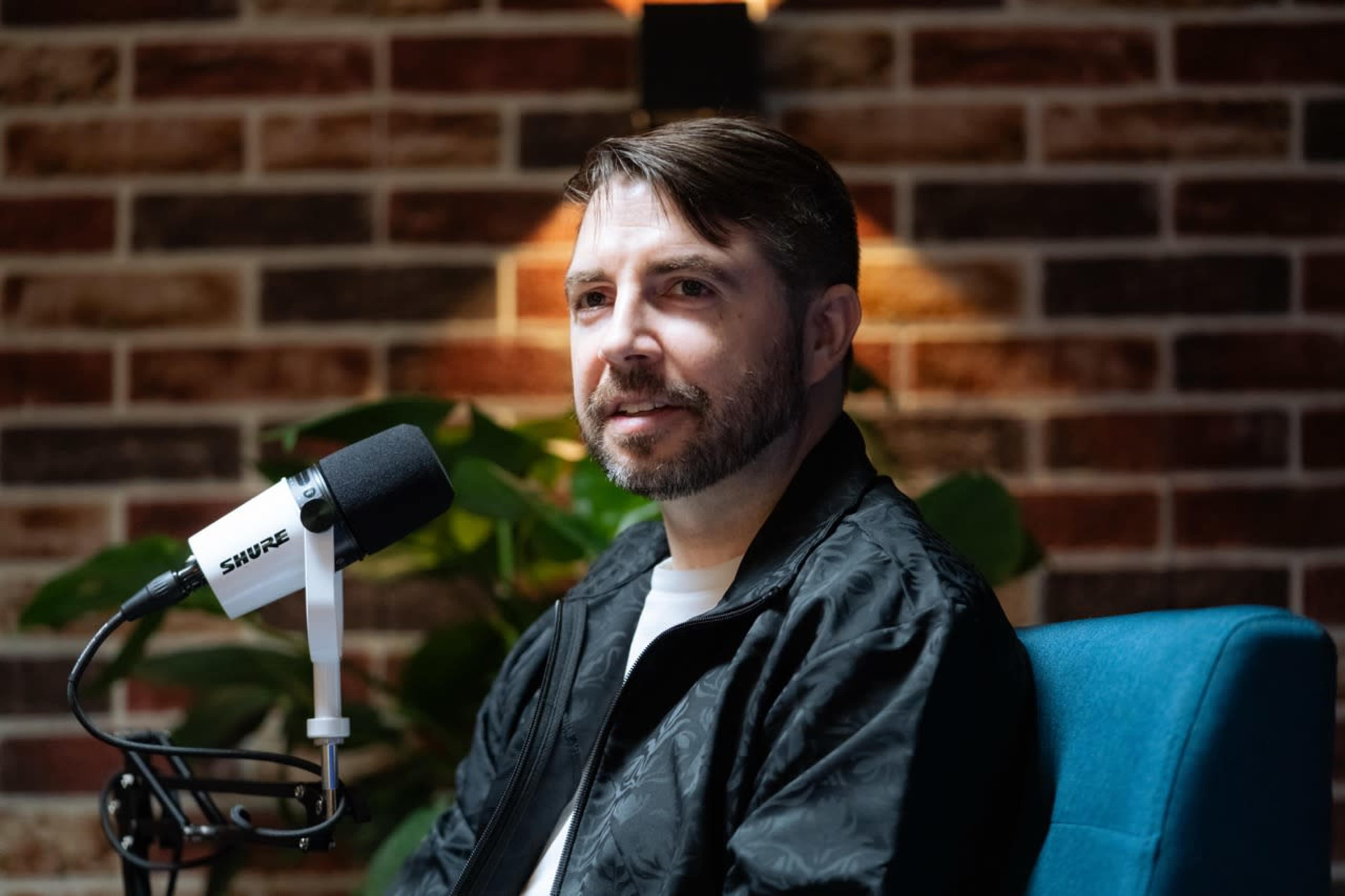 A man with a beard is sitting in a blue chair, speaking into a microphone against a brick wall with indoor plants.