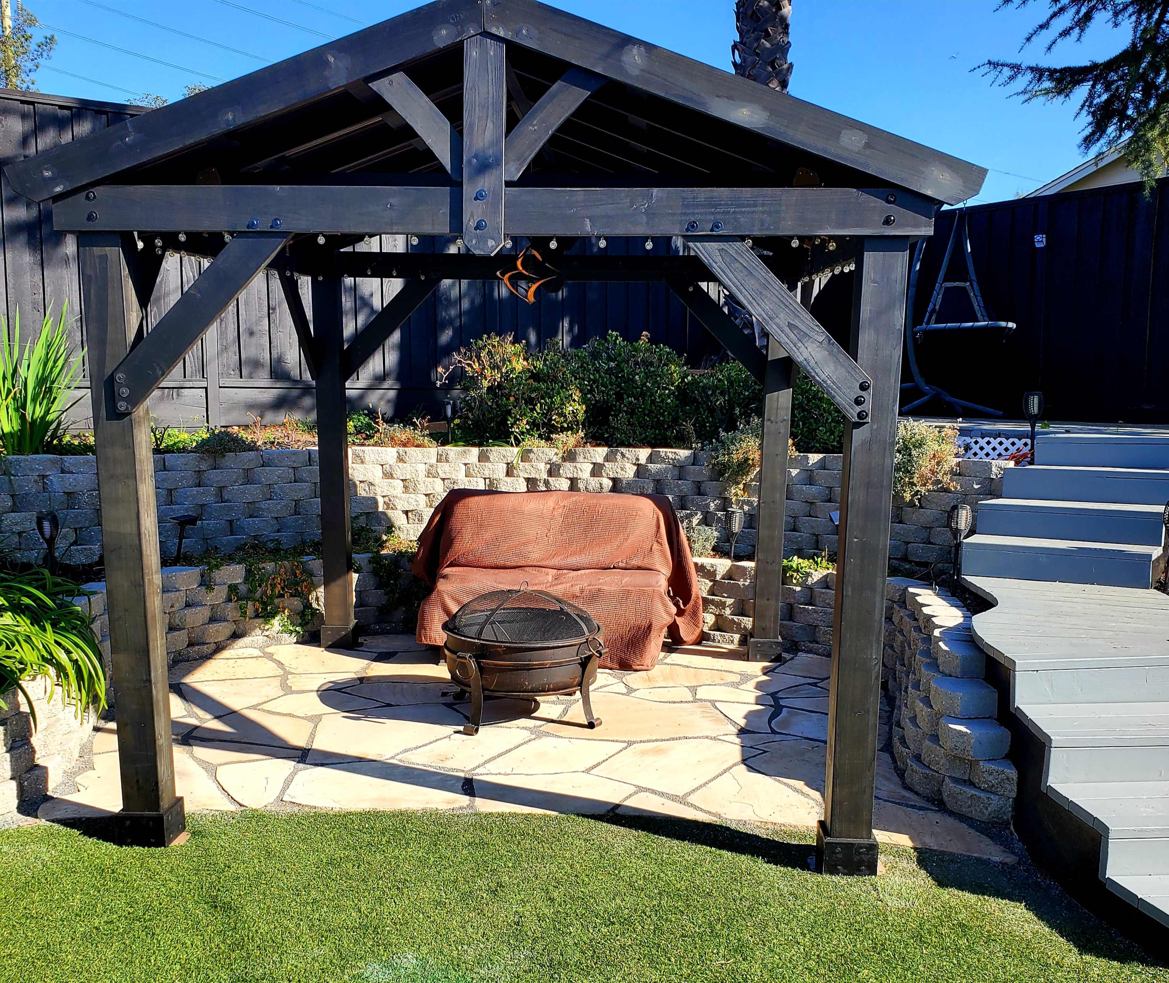 A black wooden gazebo with a stone floor, featuring a bench covered with a brown blanket and a fire pit in front of it, set in a landscaped garden area.