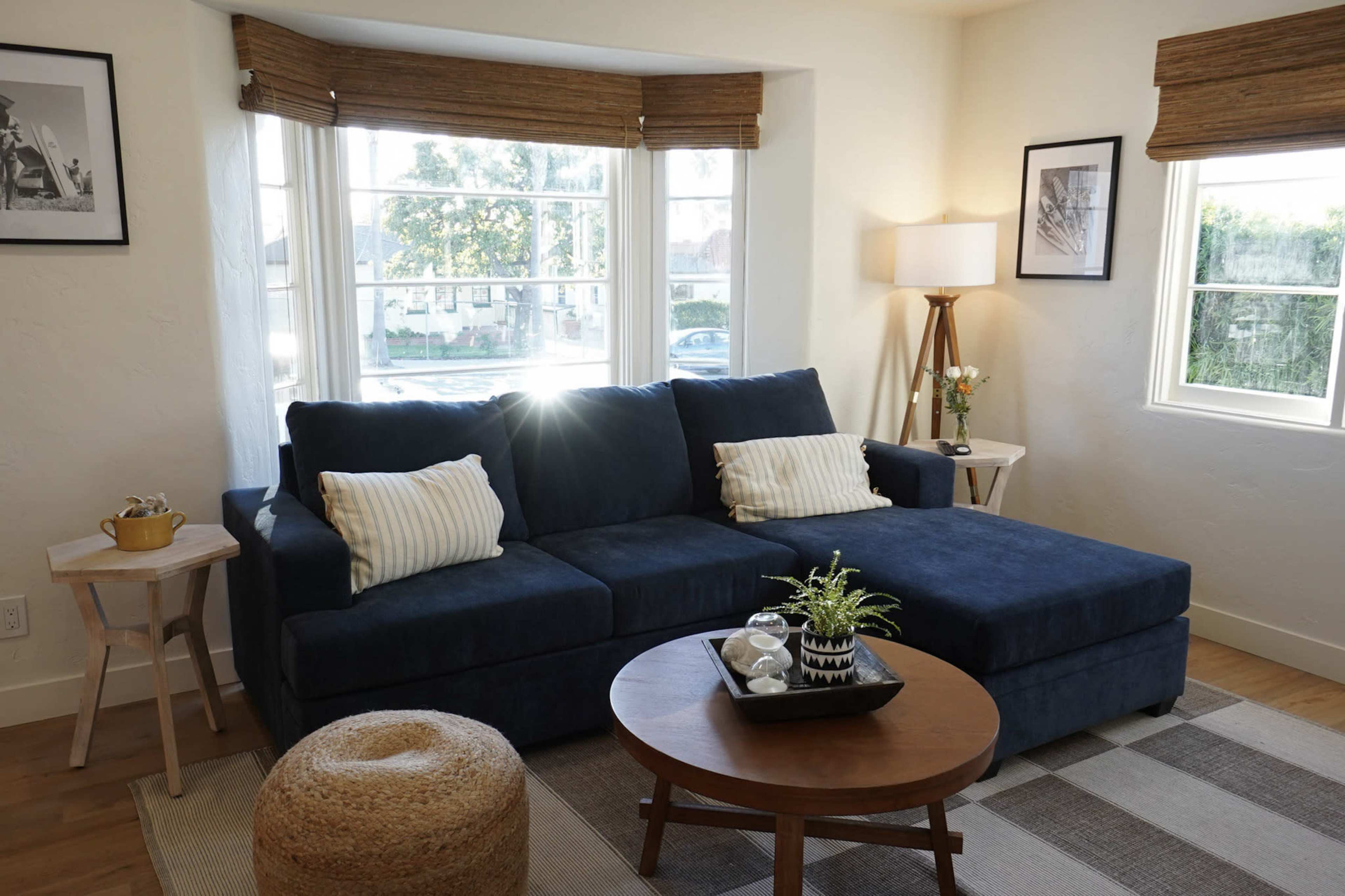 A blue sectional sofa is positioned in a bright living room with large windows, accompanied by a round coffee table and a floor lamp.
