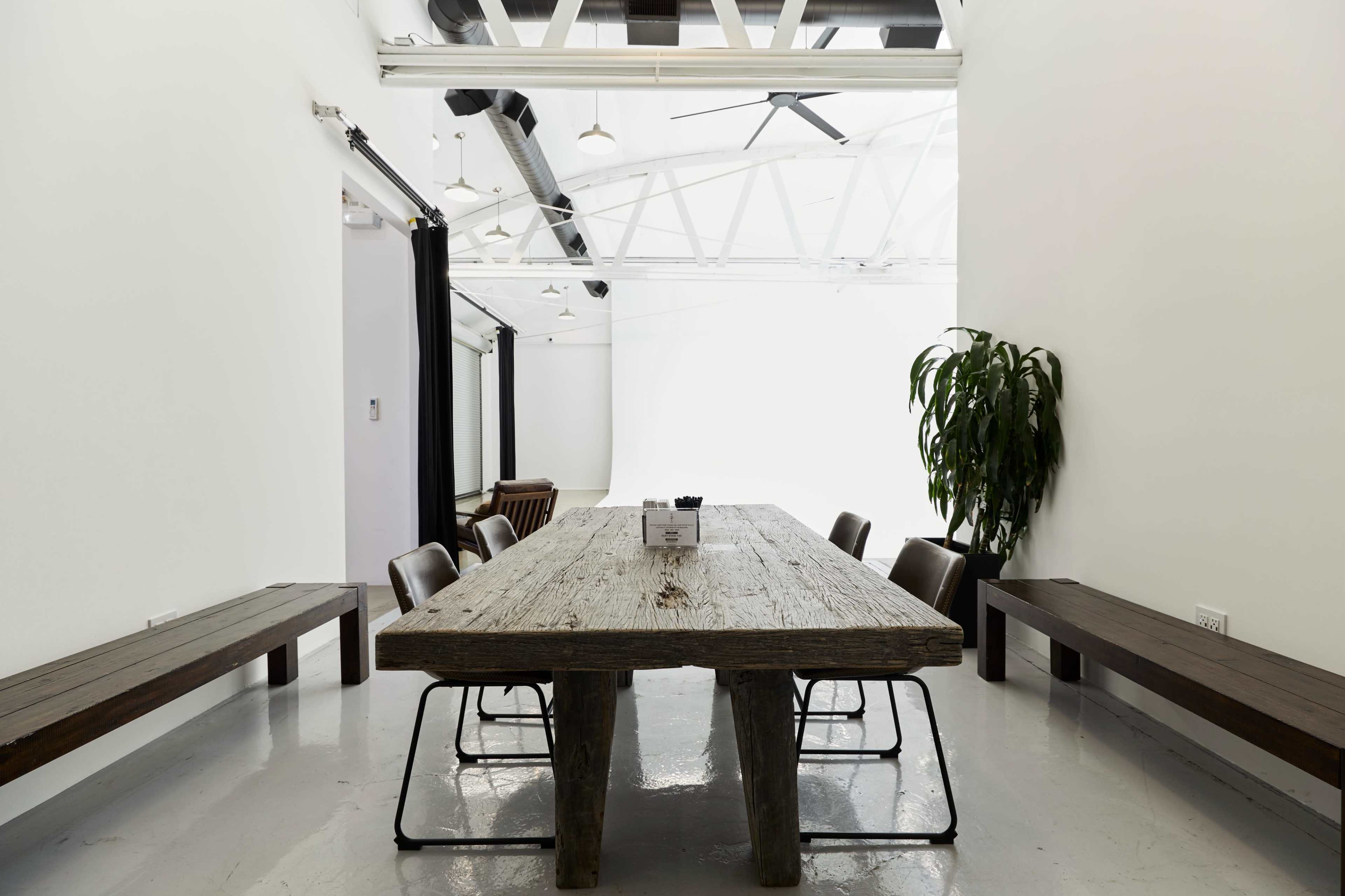 A large wooden table is positioned in a well-lit meeting room, accompanied by several chairs and a potted plant in the corner.