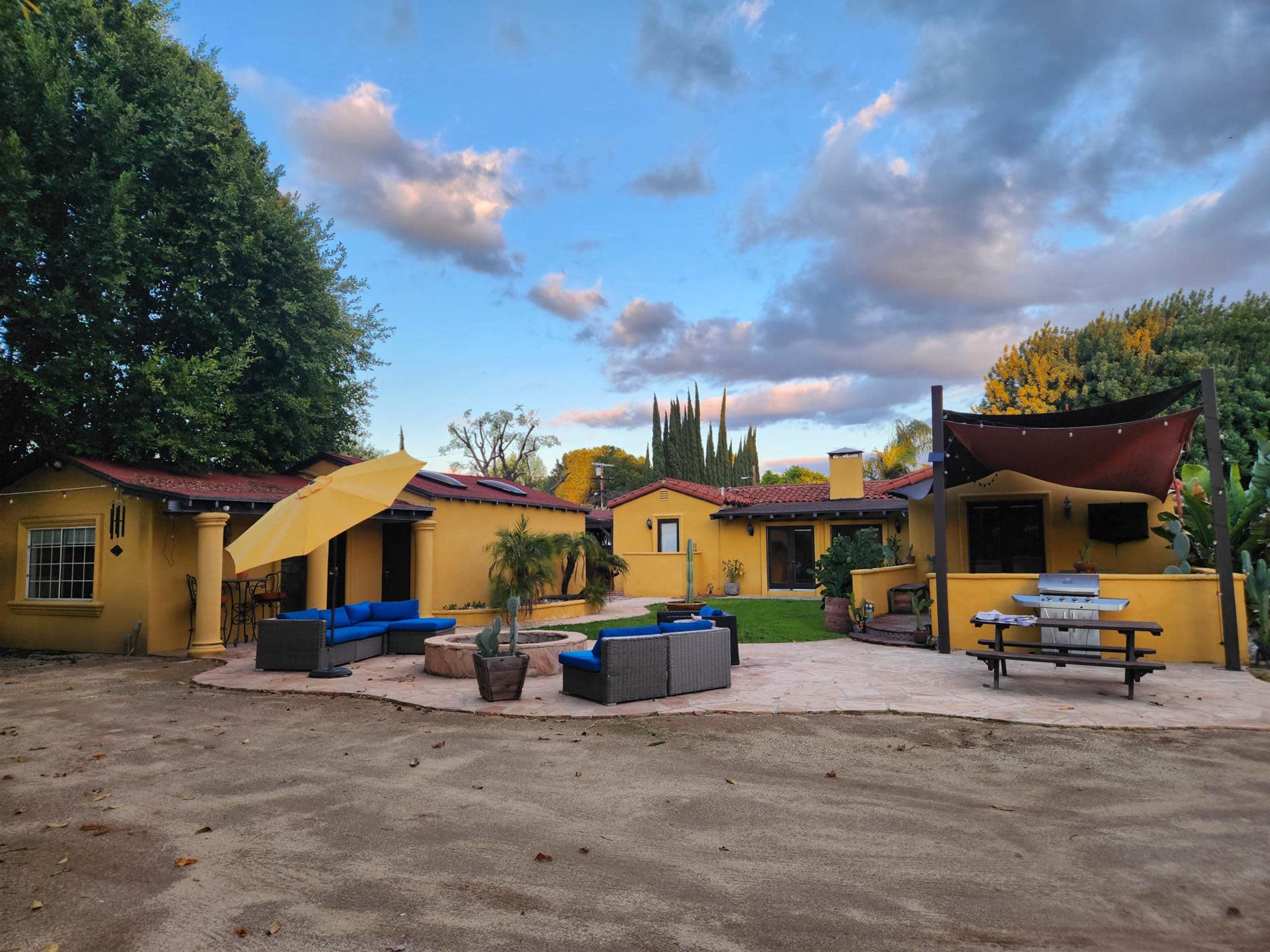 A yellow adobe-style house with a courtyard featuring outdoor seating, an umbrella, and surrounding trees under a cloudy sky.