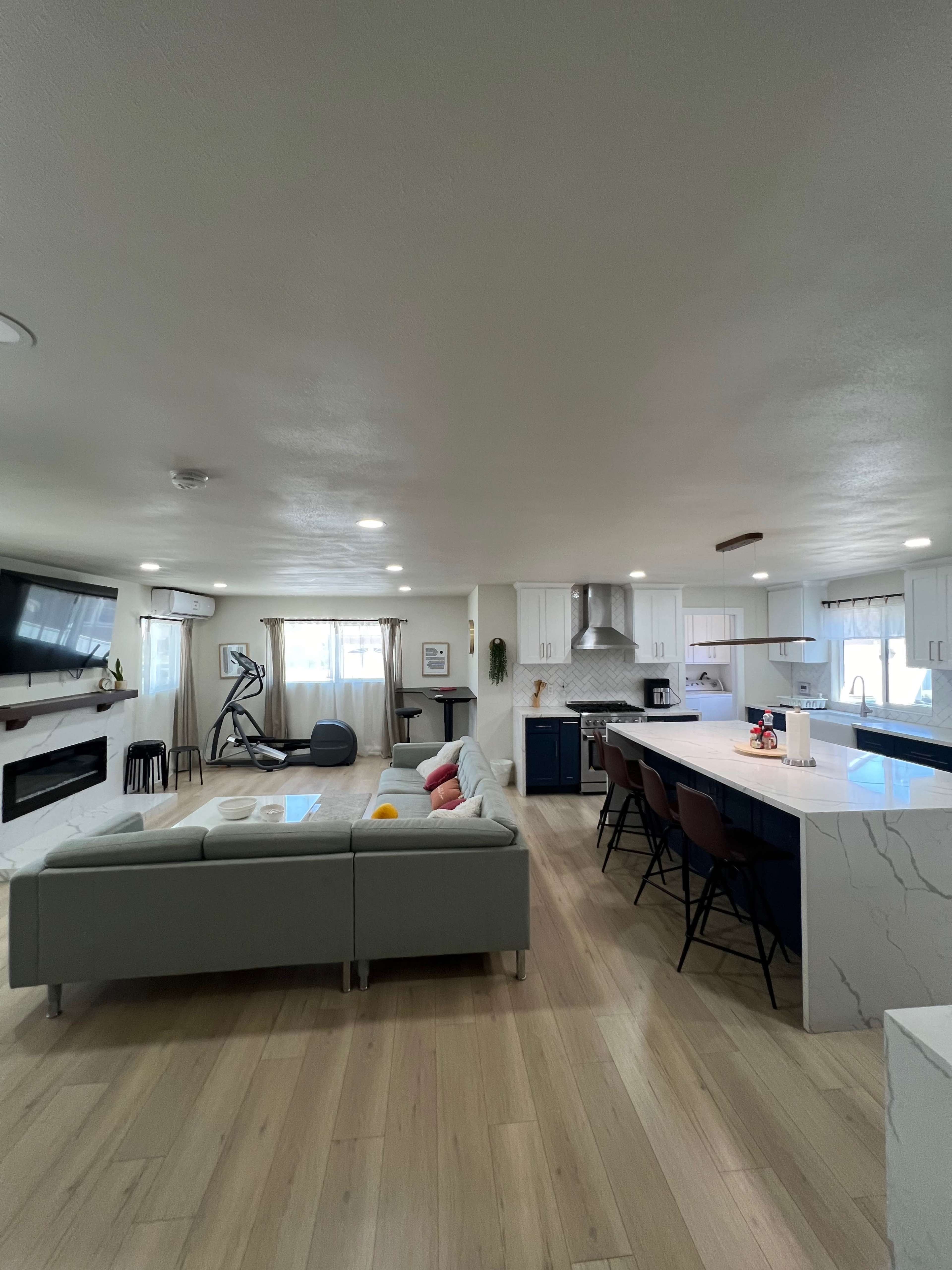 A modern open-concept kitchen and living area featuring a light-colored sofa, a large island with bar stools, and a wall-mounted television above a fireplace.