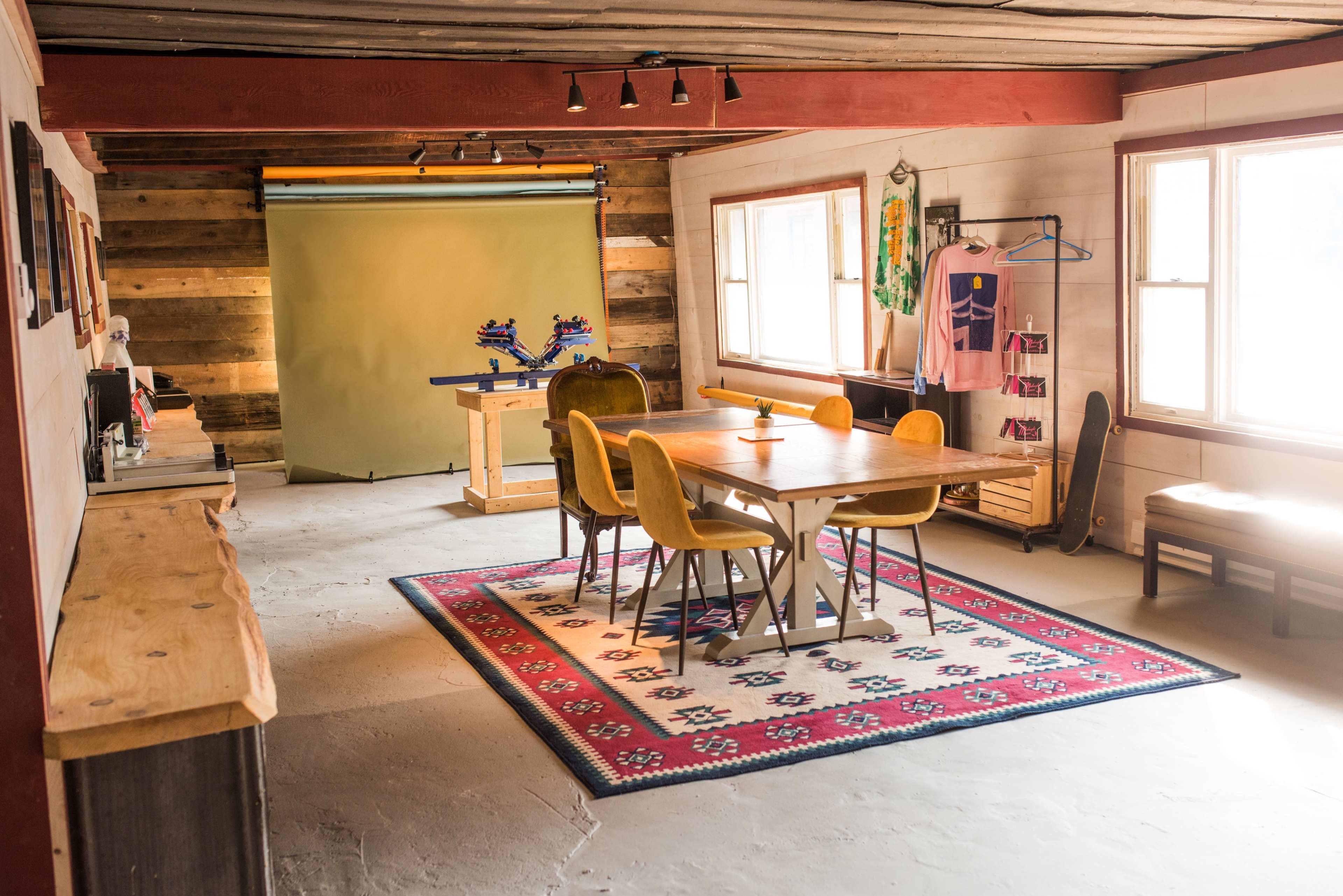 A spacious room with a wooden table surrounded by yellow chairs, a light-colored rug, and a backdrop for photography in the background.